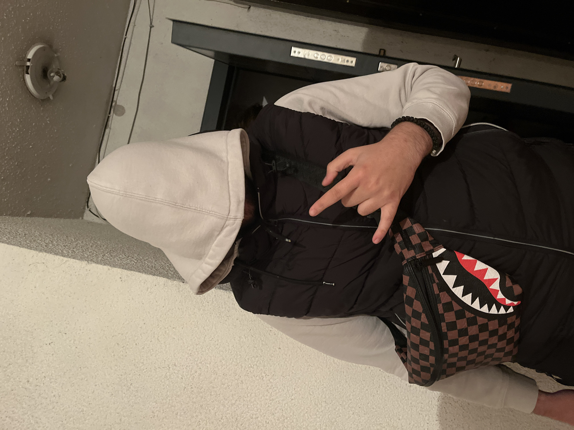 A person in a white hoodie and black vest is standing indoors, near what appears to be an elevator. The person is wearing a brown and black checkered bag with a shark's mouth design. The location in Burgas, Bulgaria is not identifiable from the image.