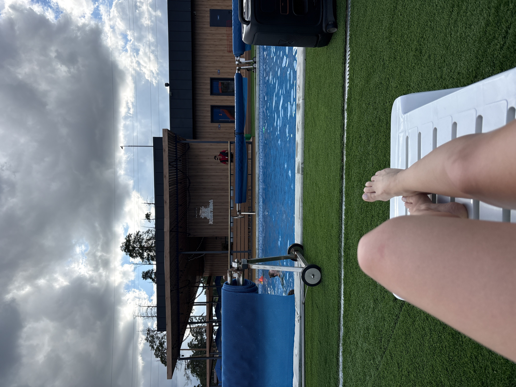 The image depicts an outdoor scene, likely in Votkinsk, Russia, judging by the Cyrillic text "ТОЧКА ТЕПЛА" (meaning "point of warmth") visible on a building near a swimming pool. A person is lounging on a white plastic chaise lounge at the pool's edge, with their bare legs and feet in the foreground. The pool is filled with blue water and has a blue pool cover partially rolled up on a wheeled mechanism. Adjacent to the pool is a strip of artificial green turf bordered by a white line. In the background is a two-story building with a dark roof and wood siding, potentially a pool house or recreation center. A person wearing red clothing is visible in the background. The sky is cloudy, and trees are visible beyond the building. Overhead power lines are present.
