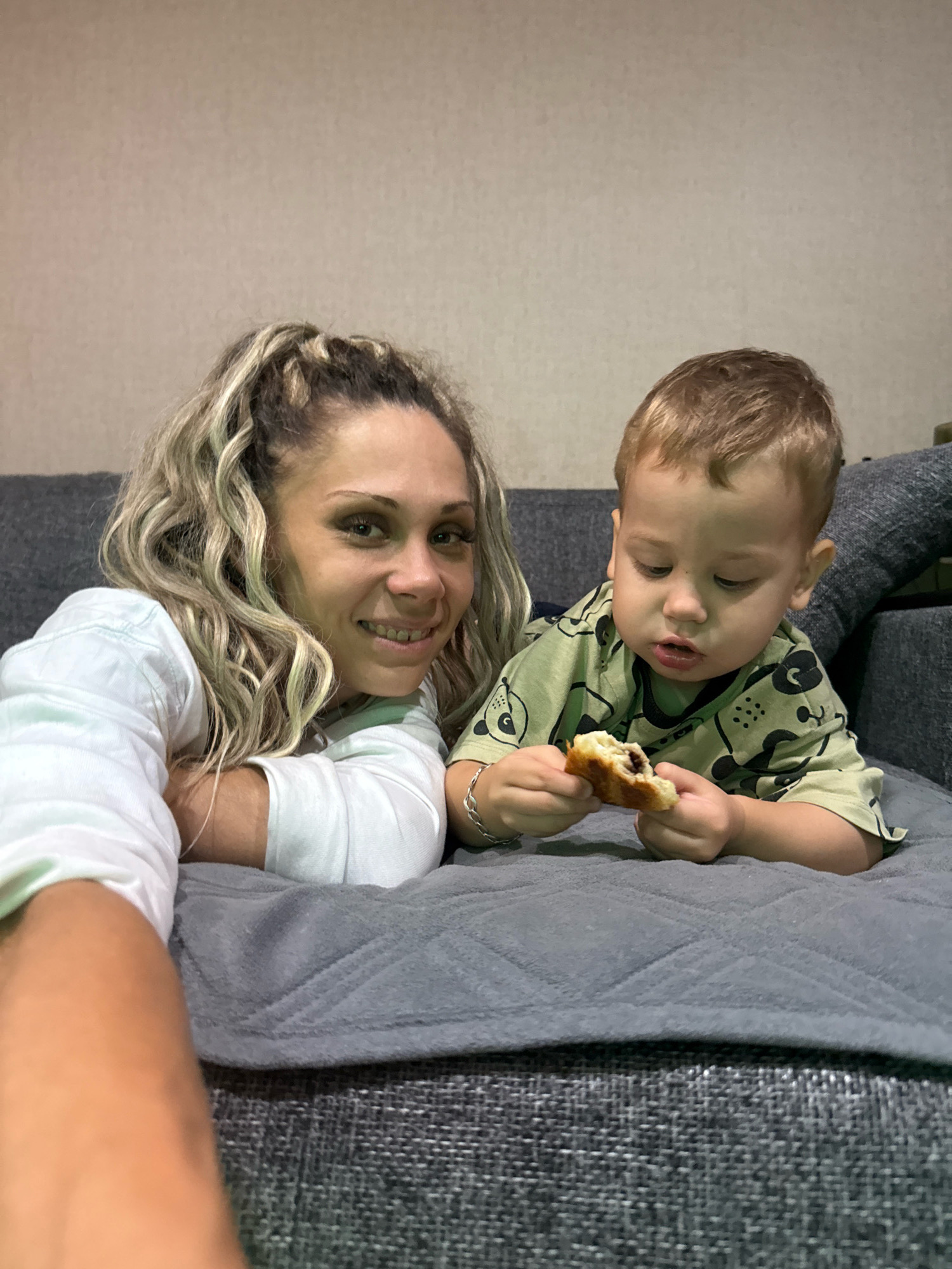 In this medium shot photograph, a woman and a young child are positioned on a gray sofa, both looking down at something. The woman, on the left, has blonde, curly hair pulled back in a ponytail and is wearing a white long-sleeved shirt. She is leaning on her forearms, looking towards the camera with a slight smile and her teeth are visible, revealing braces. The child, on the right, has light brown hair and is wearing a green t-shirt with a bear print. The child is holding a piece of pastry or bread in both hands, appearing to examine or eat it. The background consists of a textured wall. The setting appears to be an indoor environment. The image does not contain any specific landmarks or features that would allow for definitive identification of the location within Rusanovo, Russia.