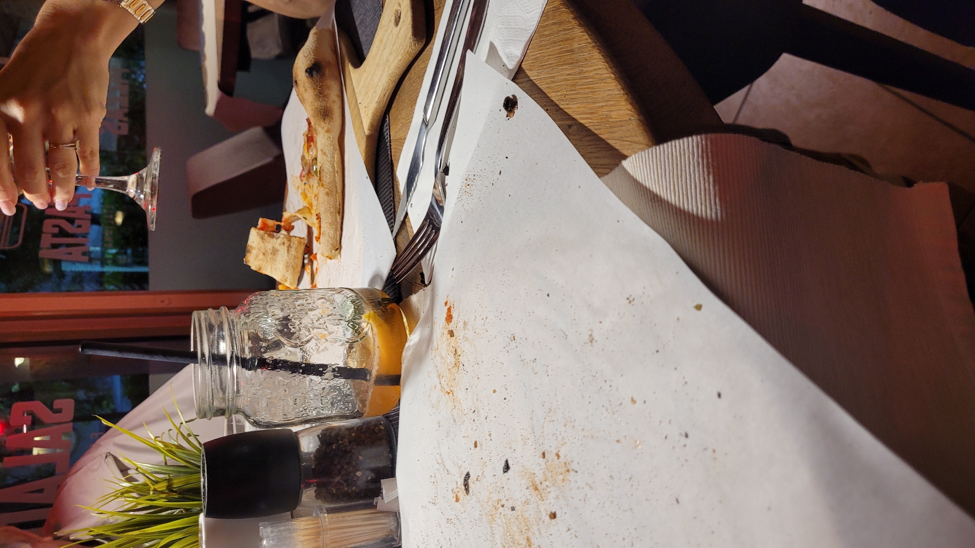 The image depicts a casual dining scene, likely in a restaurant or pizzeria, captured during the evening or night. The perspective is low, looking across a wooden table covered with white paper, which is visibly soiled with food remnants, suggesting a meal that has recently concluded or is in progress.
In the left foreground, a hand, adorned with a ring and a metal watch, holds a wine glass containing a small amount of red liquid. To the right, a clear mason jar with a black straw holds a yellow beverage. Adjacent to the jar are a small artificial green plant in a black pot and a clear spice grinder filled with black peppercorns.
On the paper-covered table, there are visible remains of a pizza, including crusts and scattered toppings. A wooden cutting board and metal cutlery (a fork and a knife) are placed near the pizza remnants. The paper is stained with crumbs and sauce, indicating active dining.
The background is dimly lit and out of focus, revealing reflections in a large glass surface, possibly a window or door. The text "ATZAM" is visible in reverse on this surface, strongly suggesting the word "PIZZA" spelled backward, which helps identify the establishment's cuisine. The artificial lighting and reflections confirm the nighttime setting. The overall atmosphere appears relaxed, consistent with a post-meal or ongoing dinner at a casual eatery in Varna, Bulgaria.