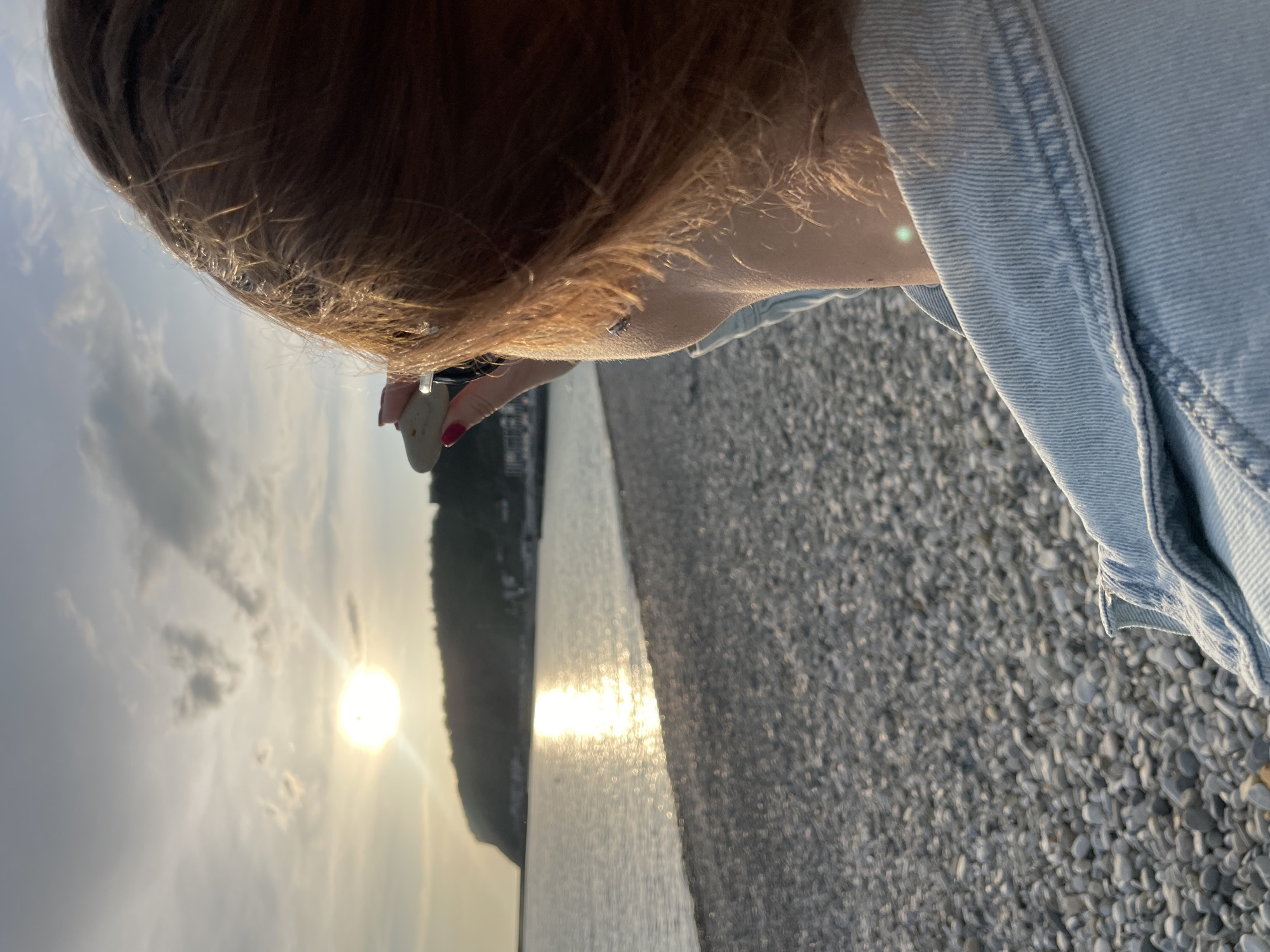 The image shows a person on a pebble beach, possibly in Olginka, Russia. The person is wearing a light blue denim jacket and holding a small, smooth stone. The sun is setting over the water, reflecting brightly on the surface. A forested hill is visible in the distance. The ground is covered with small gray pebbles.