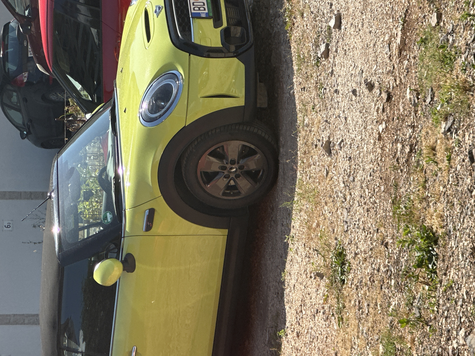 A bright lime-green Mini Cooper convertible is parked outdoors on a gravel and dirt surface, facing left in the image. The front right side of the car is prominently featured, showcasing its distinctive round headlight with a chrome bezel, black grille, and a multi-spoke wheel with a "MINI" logo. An "S" badge is visible on the side vent near the front wheel.
Behind the Mini, parts of a red car and a dark grey or black car are visible, suggesting a row of parked vehicles. The ground is covered with light-colored gravel, small stones, and patches of dry grass and sparse green vegetation.
The weather appears sunny and clear, indicated by the bright lighting and visible shadows. On the front license plate of the Mini Cooper, the letters "BD" are clearly visible, confirming the location as Budva, Montenegro. Other characters, possibly "112KE," follow the "BD" and there is a blue European-style strip on the left of the plate.
Inside the Mini's windshield, a circular green sticker is discernible. In the background, to the left of the Mini, part of a light-colored building or wall is visible, with the characters "6a" marked on it. The car appears to be parked, and no human activity is visible.