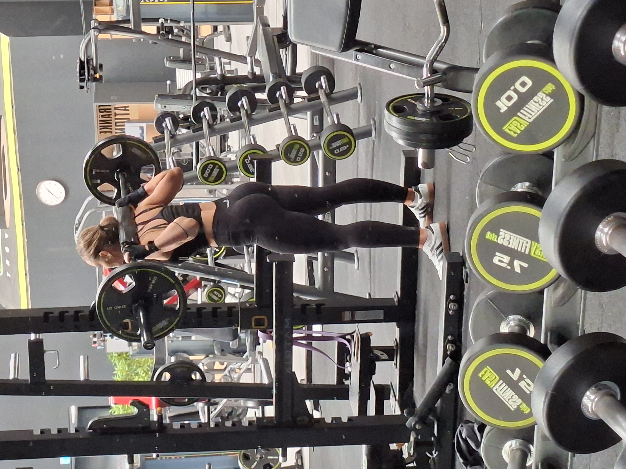 A woman is performing a barbell squat in a gym. She is wearing black workout leggings, a black sports bra, and black gloves. She has her hair pulled back in a bun. She is positioned in front of a squat rack, and a barbell with weight plates is across her upper back. The gym appears to be well-equipped with various weight machines, dumbbells, and barbells. The overall environment is typical of an indoor fitness facility. The location is identified as Baden-Baden, Germany.