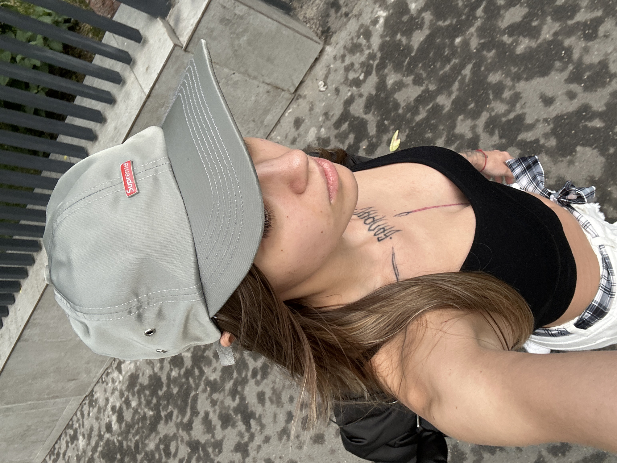 A young woman wearing a gray Supreme baseball cap, a black tank top, and white and black plaid shorts stands outdoors. Her brown hair is pulled back, and she has tattoos on her chest and neck. She is holding a black jacket over her left arm. The ground beneath her is wet and appears to be asphalt. Behind her is a gray concrete wall and a dark metal fence. There are no other people visible in the image.