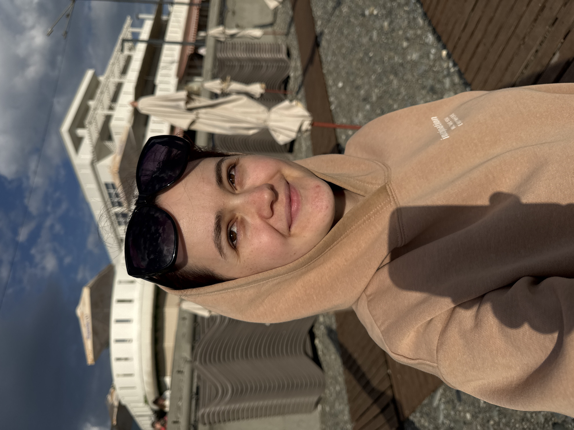 A selfie of a woman with dark hair and sunglasses, wearing a beige hoodie. She is smiling slightly. The background includes a multi-story white building with closed umbrellas, likely at a beach or resort. The ground is a mix of gray gravel and wooden decking. The photo was taken in Sochi, Russia, based on the typical Black Sea resort architecture and landscape.