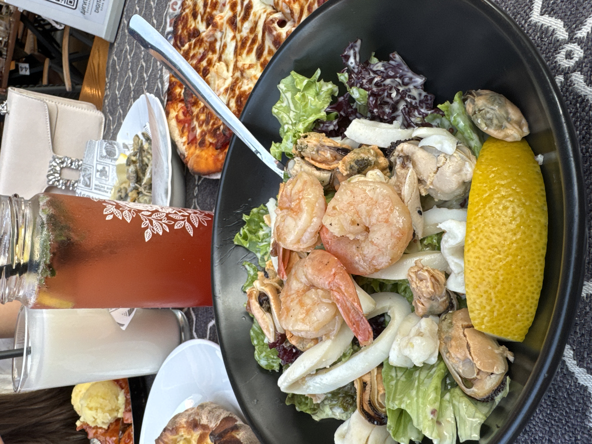 A close-up shot captures a meal laid out on a patterned tablecloth. Dominating the foreground is a black bowl filled with a seafood salad. The salad features a medley of ingredients including large pink shrimp, white calamari rings, mussels in their shells, and leafy greens, all coated in a creamy dressing. A bright yellow lemon wedge rests at the bottom of the bowl. To the right of the salad, a slice of pizza with melted cheese and red sauce is partially visible. In the background, a tall glass contains a light-colored beverage, possibly milk or a smoothie, with a black straw. Next to it, a glass jar holds a vibrant red drink with mint leaves and ice, also with a straw. Behind the drinks, a cream-colored handbag with a distinctive silver clasp is placed on the table. A small plate with what appears to be fried seafood sits to the left of the drinks. Further in the background, glimpses of outdoor seating and furniture suggest a restaurant setting. The overall scene evokes a leisurely meal outdoors, likely in a city known for its coastal cuisine.