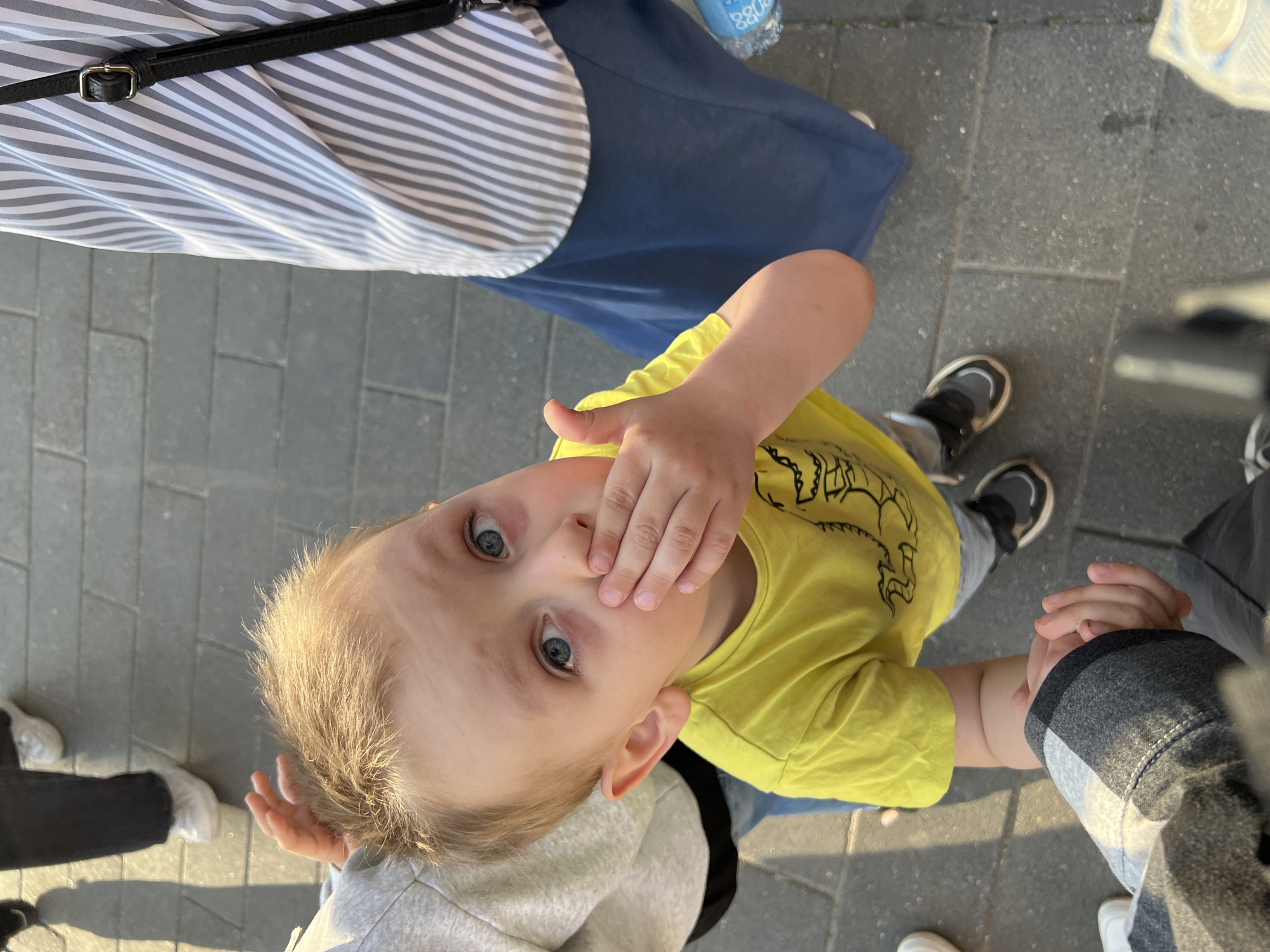 This overhead shot, taken in Mytishchi, Russia, captures a young child looking up at the camera in an outdoor, paved urban environment. The child, with fair hair and wide blue eyes, is wearing a bright yellow t-shirt and grey and black sneakers. One of their hands is covering their mouth, conveying a sense of shyness, surprise, or curiosity, while their other hand is held by an adult's hand, visible with a grey sleeve in the bottom right.
Several adults' body parts are visible around the child. In the upper left, a person is wearing a grey and white striped top, dark blue attire (likely a skirt or trousers), and has a black strap with a silver buckle across their body. Another pair of legs in black trousers and white sneakers is visible in the lower left, casting a shadow on the dark grey paving stones that form the ground.
The lighting suggests it is either late afternoon or early evening, characterized by the noticeable shadows on the pavement. A plastic water bottle is partially visible in the upper right corner, with some indistinct text, possibly "ББО" or "880", printed on it. The image portrays a candid moment of a child's interaction and observation in a public, sunlit setting.