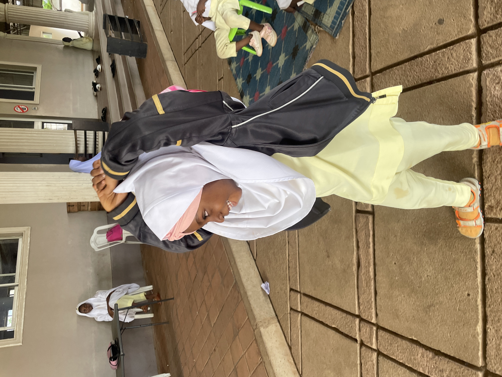 In Barakin Lerawi, Nigeria, a young girl wearing a pink hijab and a white veil is pictured from the waist down. She is wearing a yellow top and pants, and orange sandals. She is holding a black jacket with gold trim over her shoulders. In the background, another girl in a white hijab and yellow pants is sitting on a chair. Other people are visible in the background, some seated on the floor and others on chairs. The scene appears to be set in a mosque or a similar religious or community gathering place, with tiled flooring and architectural columns.