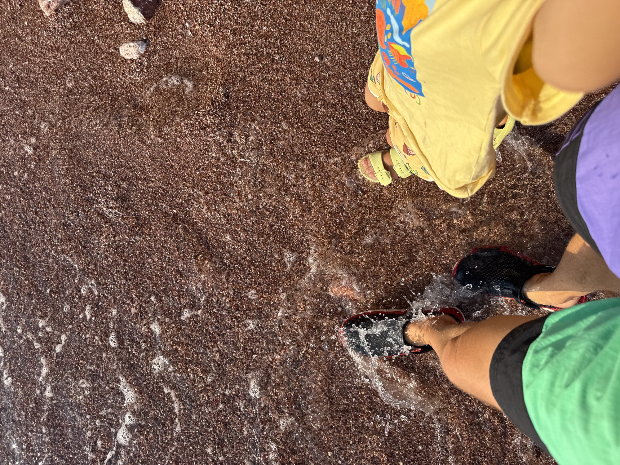 This is a POV shot looking down at two people standing in shallow water on a pebble beach. The person on the left is wearing black water shoes with red accents and bright green shorts. The person on the right is wearing yellow sandals and a yellow t-shirt with a colorful cartoon-like print. The beach is covered in small, dark red pebbles, with some small waves lapping at the shore, creating white foam. The scene appears to be on the coast of Sveti Stefan, Montenegro.