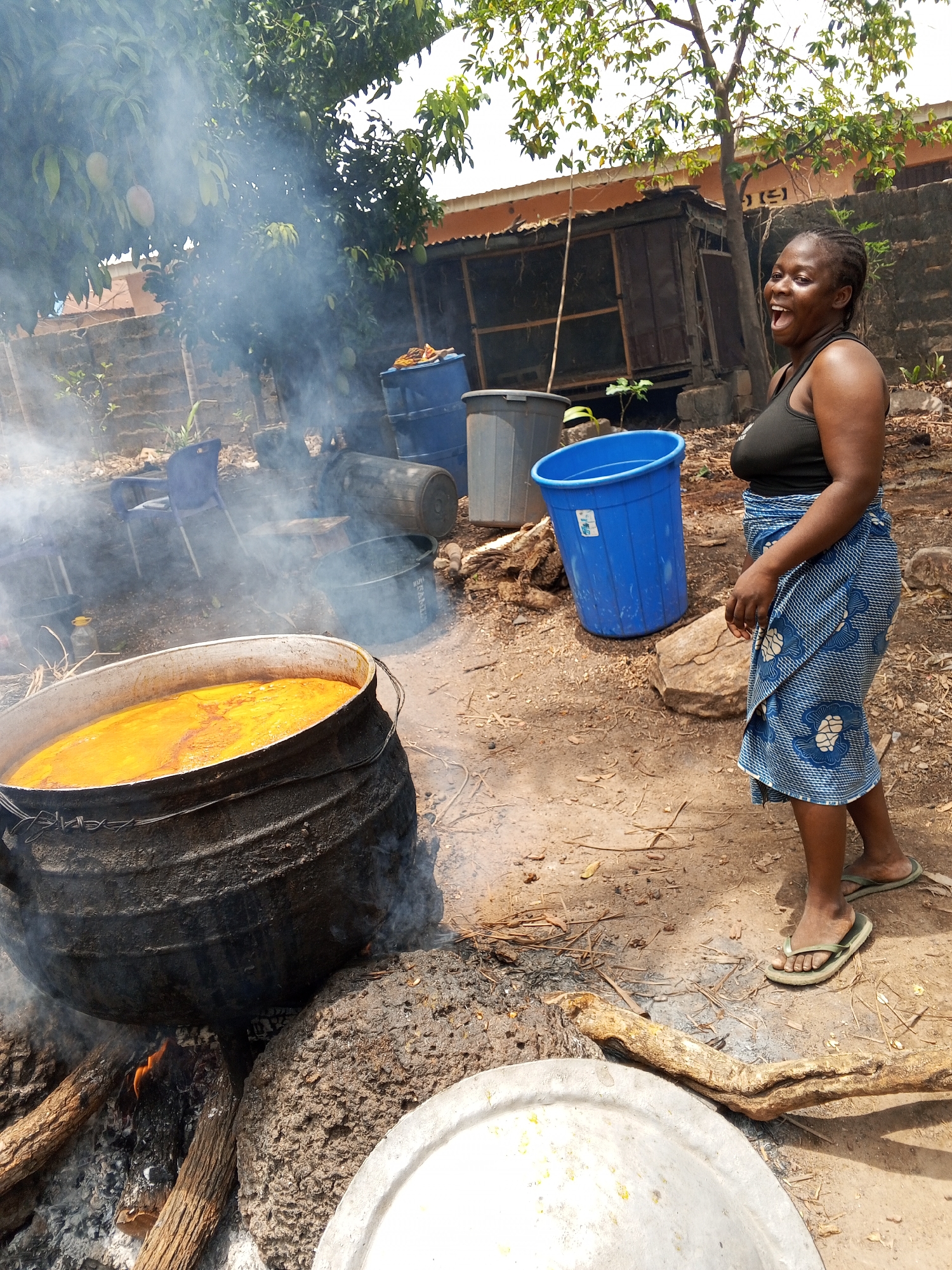The image shows a scene in Akwanga, Nigeria. A woman is standing outdoors near a large metal pot cooking over an open fire. The pot is full of a yellow liquid that appears to be boiling, with smoke emanating from the fire and pot. The woman is wearing a black tank top, a blue patterned cloth wrapped around her waist, and flip-flops. She is smiling and has her mouth open. In the background, there are trees, a makeshift wooden structure, and various containers, including blue plastic bins. The ground is dirt and scattered with leaves and twigs. There is a large metal lid on the ground near the pot.