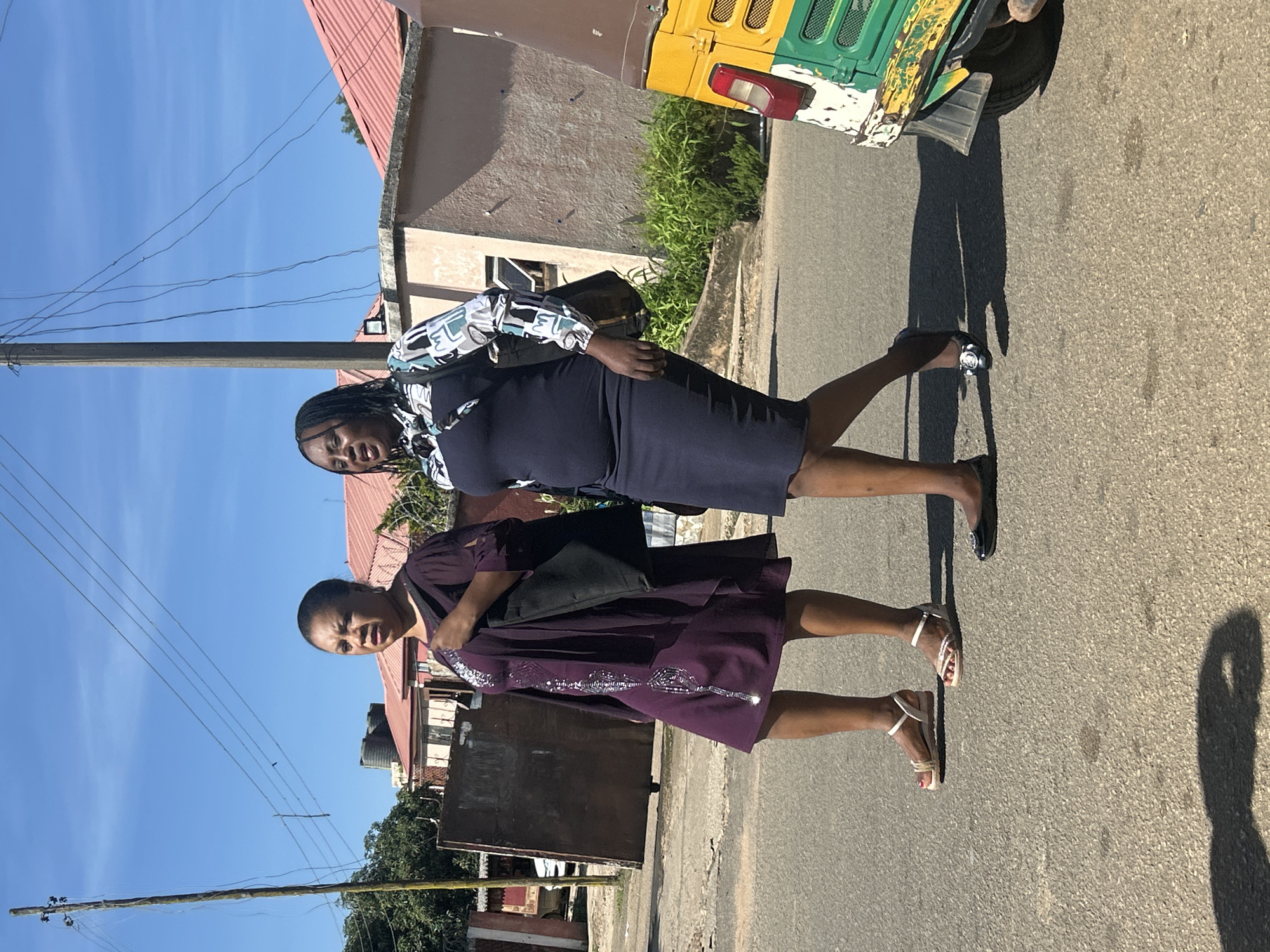 Two women are walking on a paved road under a clear blue sky in Jos, Nigeria. The woman on the left is wearing a knee-length purple dress with silver embellishments and light-colored sandals. She is carrying a black tote bag over her right shoulder. The woman on the right is wearing a dark blue dress with a patterned long-sleeved top and black ballet flats. She is also carrying a dark bag over her right shoulder. Behind the women, there are buildings with red roofs and some greenery. A utility pole with overhead wires is visible to the left. A colorful auto-rickshaw is parked on the right side of the road.