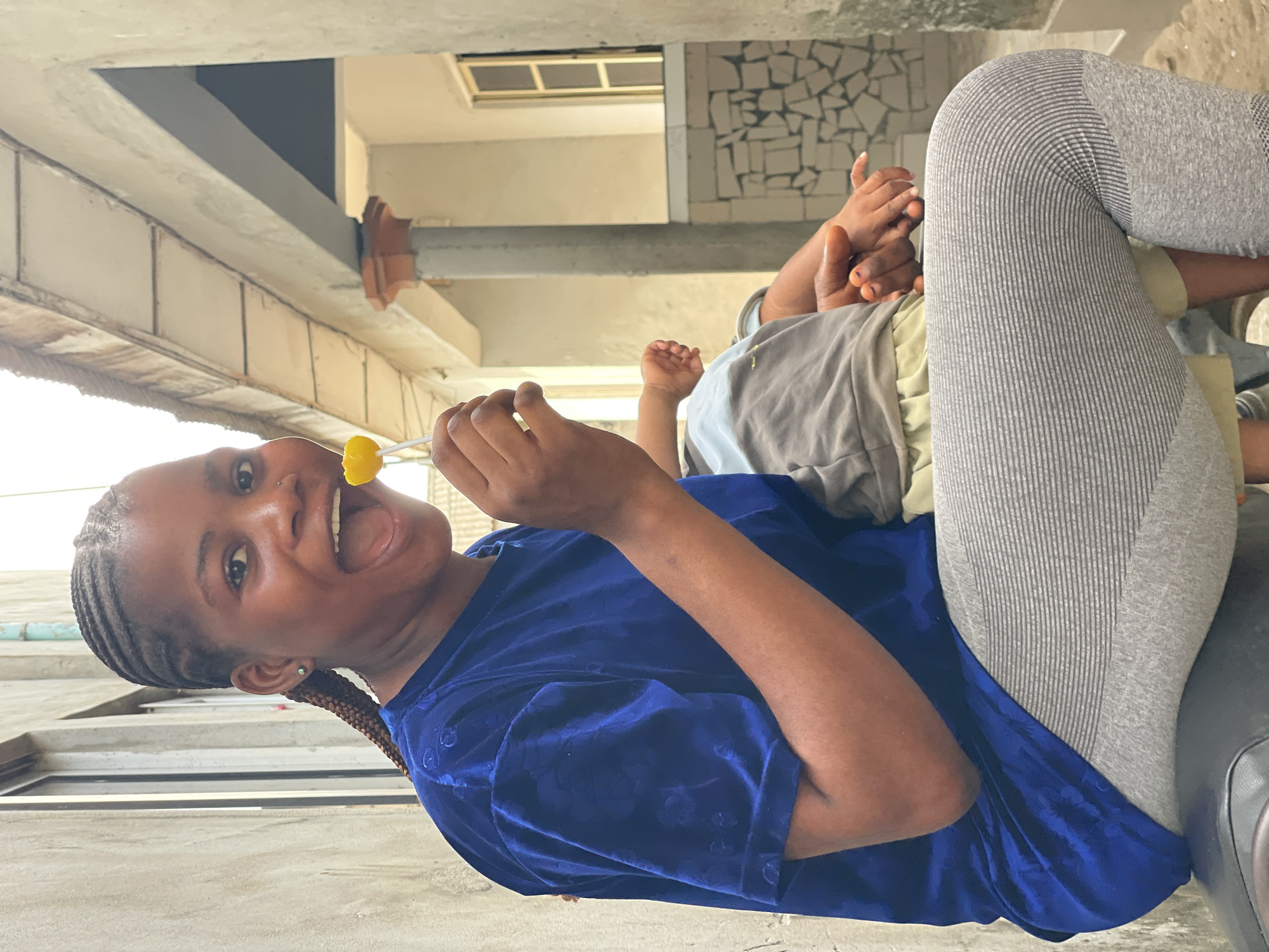 A woman in a blue shirt and gray leggings sits with a child on her lap. The woman is holding a yellow lollipop in her right hand and has a wide smile. She has braided hair pulled into a ponytail. The child is wearing a gray shirt. The background appears to be a building exterior. The image seems to be taken in an urban area of Badagry, Nigeria, as can be inferred from the visible architectural style and construction materials.