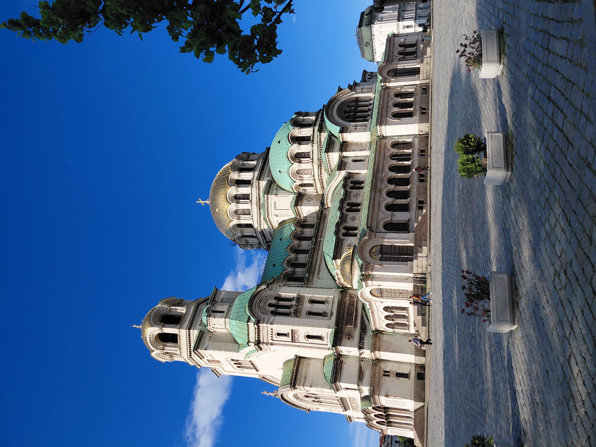 The image showcases the St. Alexander Nevsky Cathedral in Sofia, Bulgaria, under a clear, bright blue sky with a few scattered clouds. A tree branch with green leaves appears in the top left corner. The cathedral's architecture is prominent, with white walls, green-patinated domes, and golden accents. Stone paving covers the ground beside the cathedral, where there are decorative rectangular planters with some plants. Several people are walking along the cathedral's side, indicating human activity in the scene. The overall environment is urban and architectural, showcasing the cathedral as a landmark within the city.