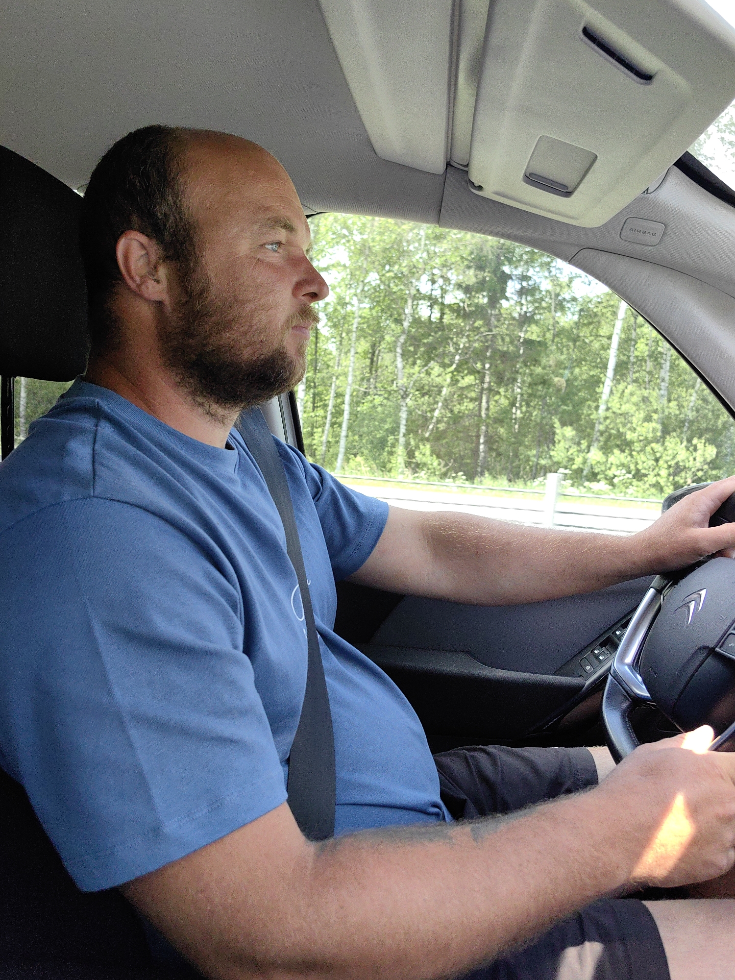The image captures a male driver operating a vehicle in Негарэльскі сельскі Савет, Belarus, during daylight. The man, with a receding hairline and a full beard, is seated in the driver's position, wearing a blue short-sleeved t-shirt and dark shorts. A black seatbelt is visible across his chest and lap. He is looking forward and slightly to the right, out the front windshield. His left hand grips the black steering wheel, which has a silver-colored spoke and a discernible Citroën emblem. Sunlight illuminates his right forearm and hand, also positioned on the steering wheel. The vehicle's interior features a light grey headliner, a light grey sun visor with a central opening, and a light grey A-pillar marked with "AIRBAG." The left door panel, visible below his arm, is dark and contains window control buttons. Outside the vehicle, a dense treeline of green deciduous trees is visible, indicative of a forested environment. A white roadside barrier or guardrail runs parallel to the road.