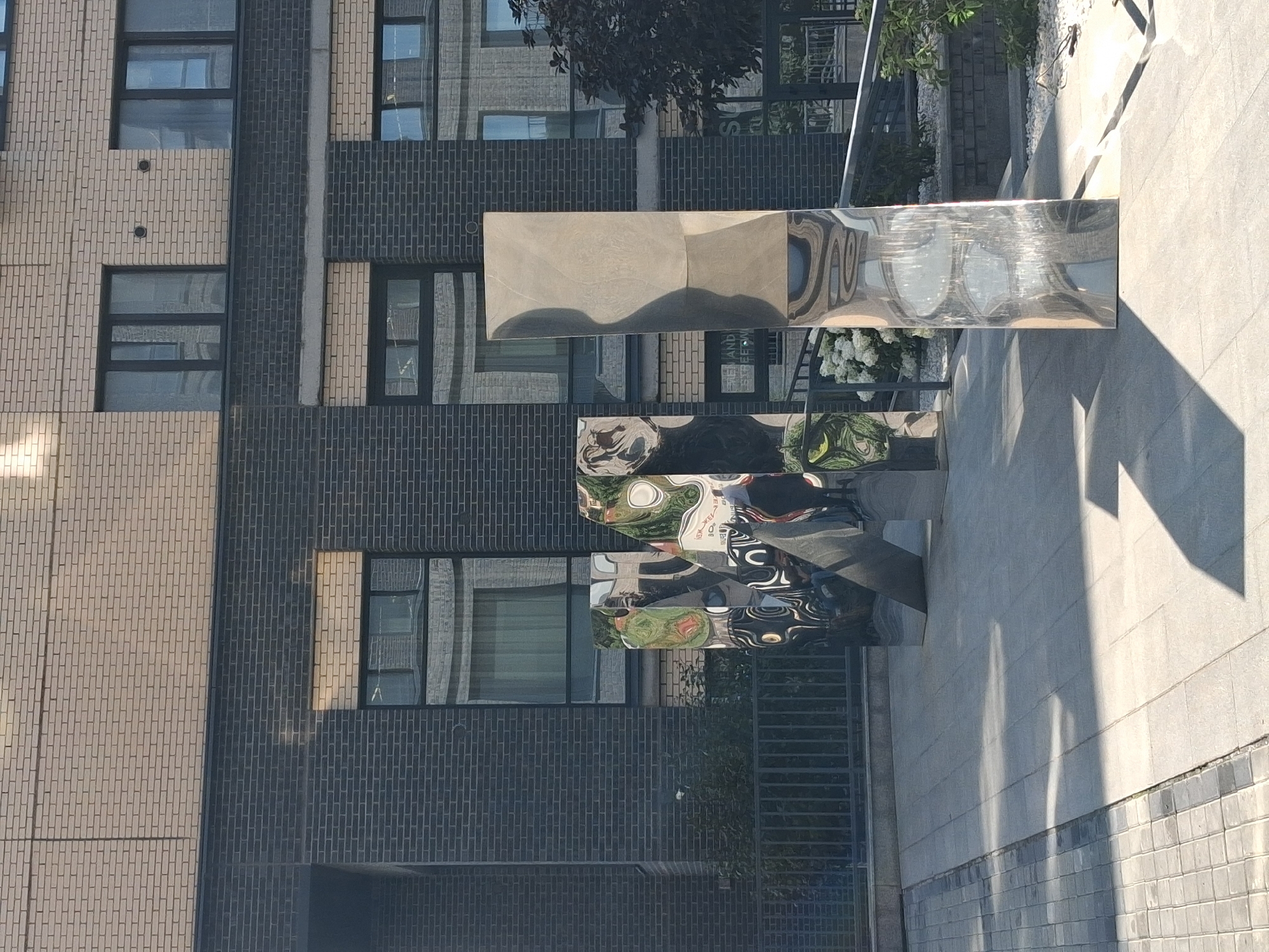 The image shows a modern building in Novosibirsk, Russia. The building has a facade of beige and dark grey brick. In front of the building are two large, abstract sculptures made of polished metal. The sculpture on the left is made up of several reflective panels forming a fragmented shape. The sculpture on the right is a single, tall, rectangular prism with a reflective surface. Both sculptures reflect the surrounding environment, including the building, sky, and vegetation. The ground in front of the building is paved with light-colored square tiles. A metal fence runs along the edge of a planted area behind the sculptures. Sunlight casts shadows on the paved area. The overall scene depicts an urban architectural setting with contemporary art installations.