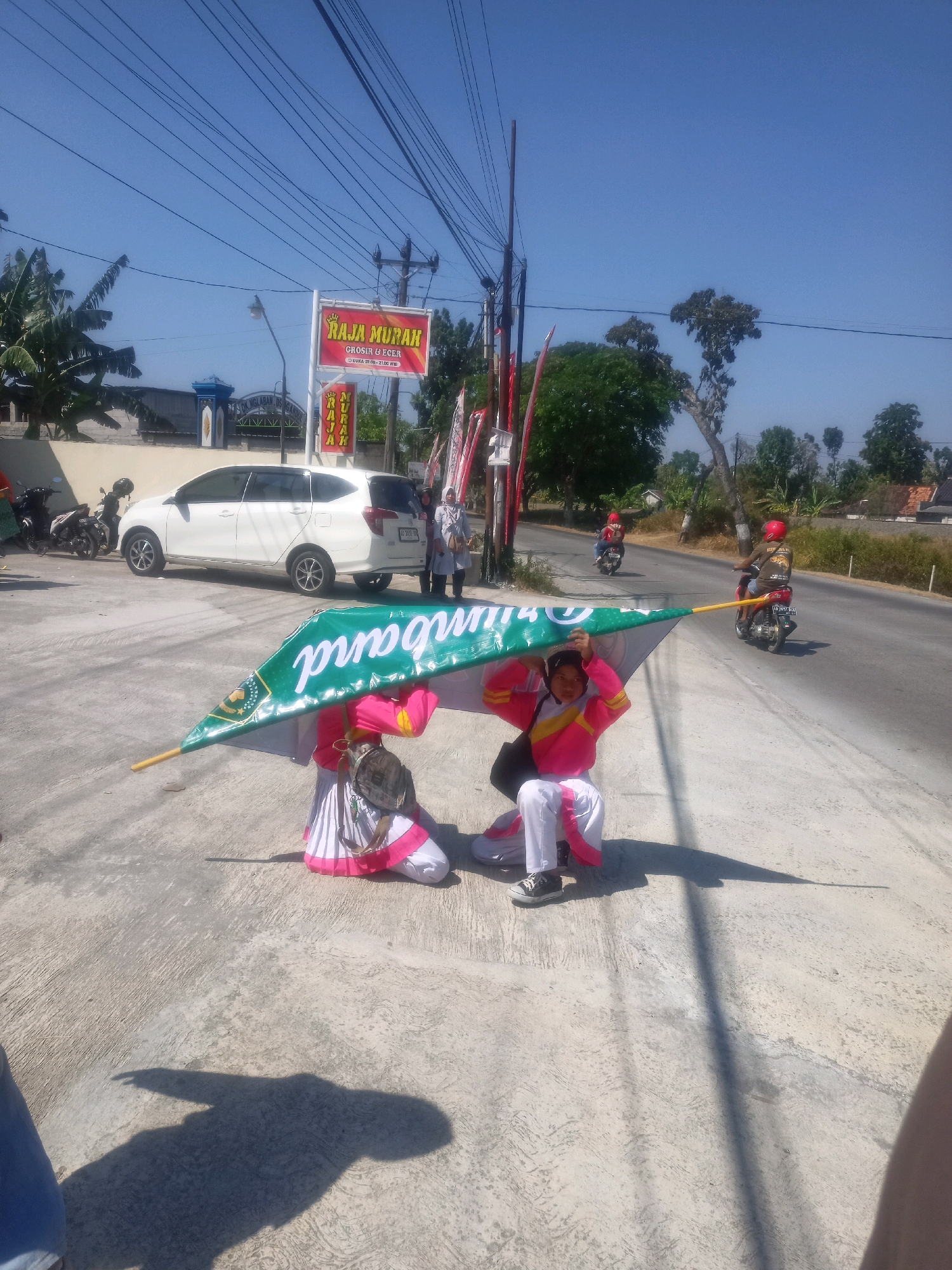 In Sumberlawang, Indonesia, under a clear blue sky and a web of power lines, two individuals dressed in matching marching band uniforms are captured mid-action. One person, in the foreground, is kneeling and holding a large green banner with white text that reads "GENTA KECANA MARCHING BAND". The banner is supported by a wooden pole extending out of frame. Their uniform consists of a pink jacket with yellow accents and a white skirt with pink trim, paired with black sneakers. They are wearing a black bag slung over their shoulder. Behind them, another person in a similar uniform is also kneeling, obscuring part of the banner. In the background, a white minivan is parked, with a few motorcycles nearby. Further down the road, two individuals are riding motorcycles, one of whom is also wearing a red helmet. The scene unfolds on a concrete surface next to a paved road lined with trees and buildings, suggesting a public space or a road in a town. A sign advertising "RAJA MURAH" (Cheap King) is visible on the left side of the image. The overall impression is a bright, sunny day, likely during a parade or public event involving the marching band.
