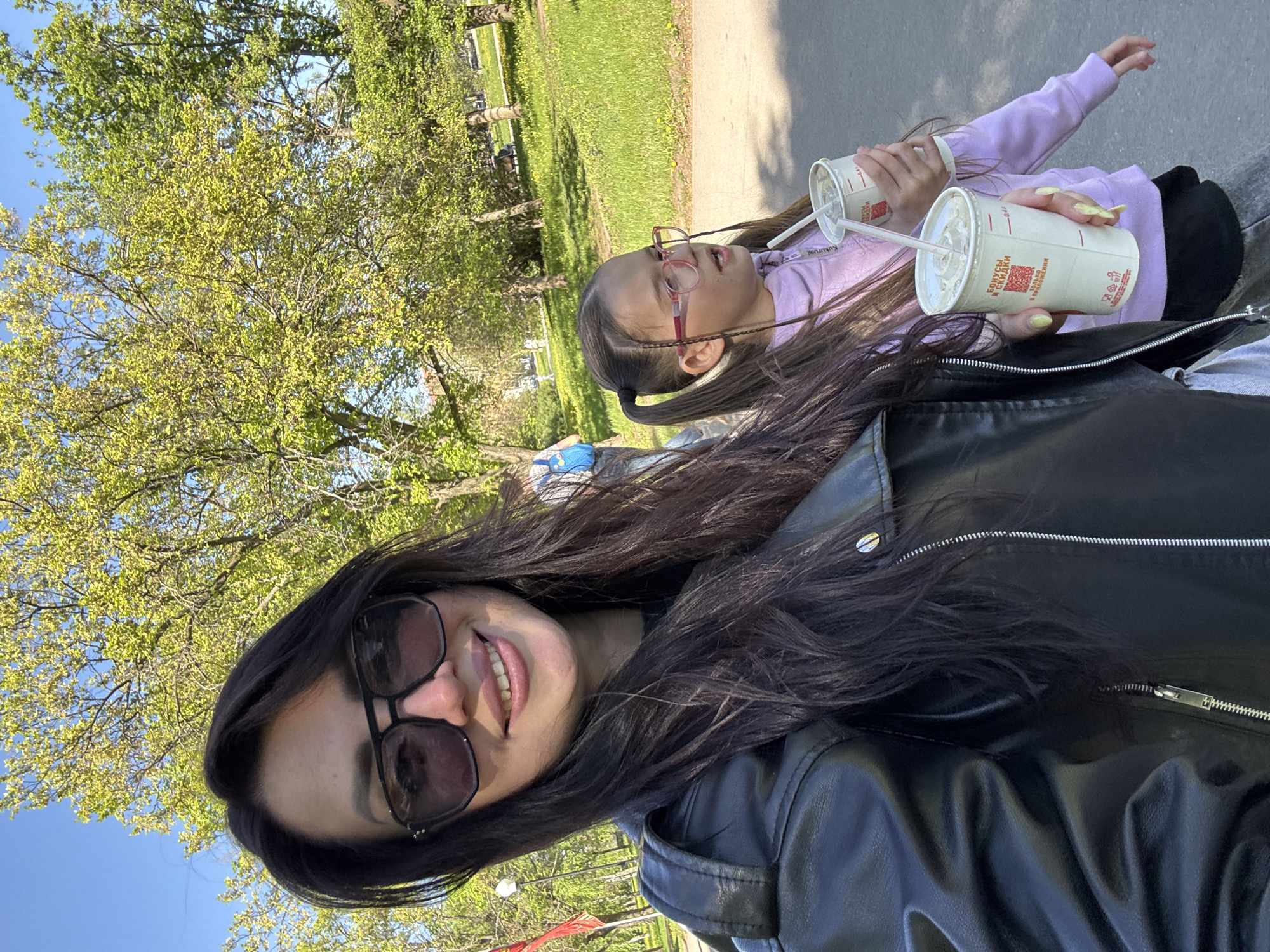 The image depicts a selfie of a woman with her child in an outdoor setting, likely a park. The woman has dark hair, is wearing sunglasses, and a black leather jacket, smiling at the camera. Beside her is a child with long brown hair, wearing glasses and a light pink sweatshirt. Both are holding paper cups with straws. The background shows green grass, trees, and a paved path. The location appears to be a park within a city.