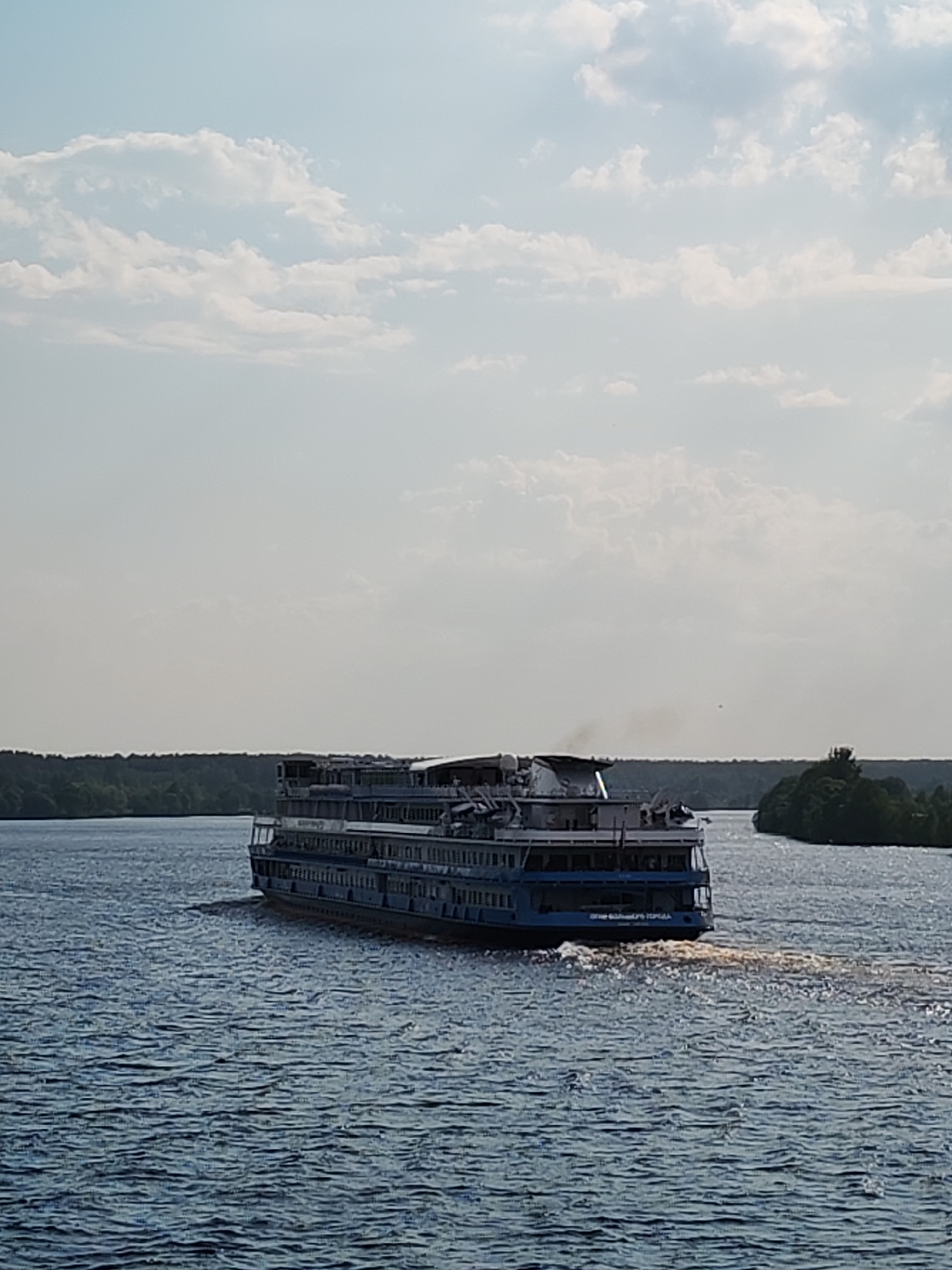 A river scene captures a large, multi-story passenger ship moving away from the viewer on rippled water, with dense, dark green tree lines on the shores in the background under a cloudy sky. The ship has a blue stripe along its lower hull and a dark section on top where a small plume of black smoke is visible. The location is the city of Unknown, Unknown.