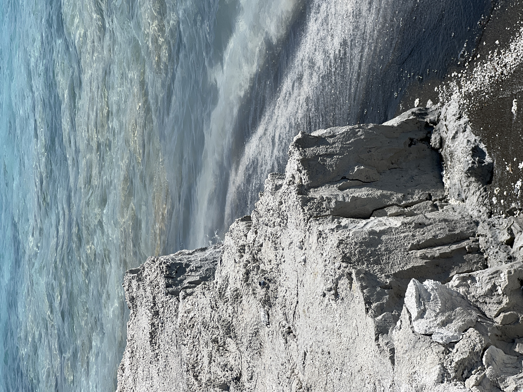 The image shows a close-up view of a light-colored, rocky cliff face meeting a body of water. The cliff is composed of rough, textured material with visible layers and cracks, appearing eroded. The water is a clear, light blue or turquoise color, with ripples on its surface suggesting movement. Towards the shore, the water becomes darker, with visible sediment or lighter-colored debris swirling near the cliff's base. The scene appears to be a natural landscape. The provided context states this is in Yeşilova, Turkey, likely referring to a unique geological formation or body of water in that region. There are no people or animals visible in the image.