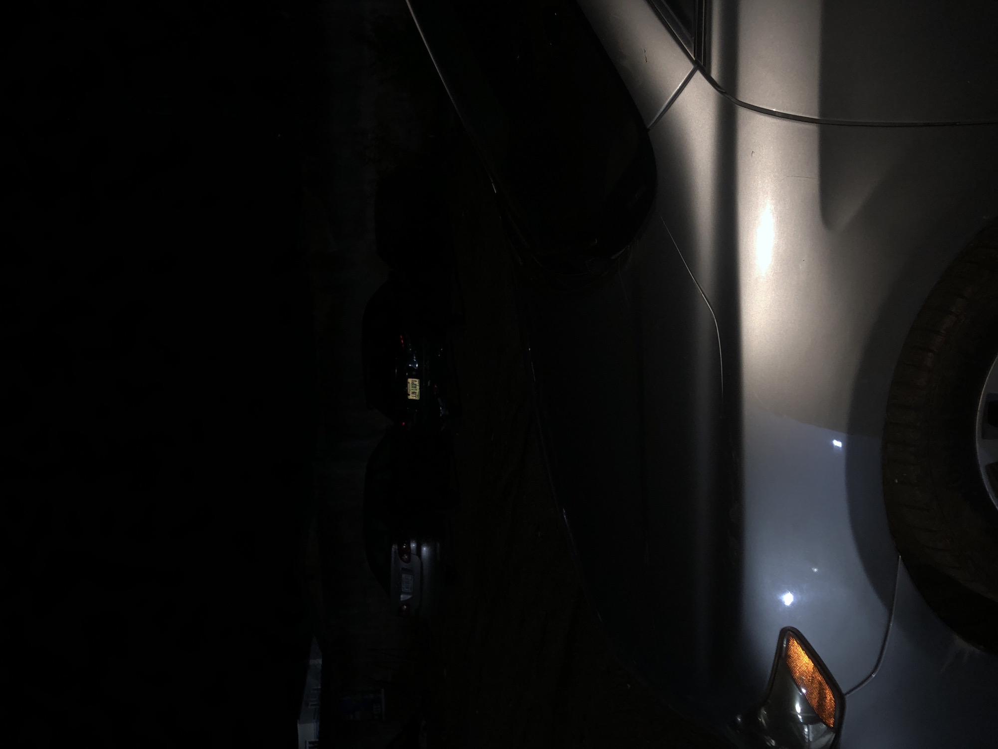 The image depicts an outdoor, low-light night scene, likely a parking area or street, in the city of Unknown, Unknown. In the immediate foreground, the front right quarter of a silver or light grey sedan is prominent, illuminated by an unseen light source originating from the left. Visible features include a portion of the headlight assembly with an amber turn signal or reflector, the front fender displaying a minor dent or crease and several superficial scuff marks, and the upper section of the front right wheel and tire. The vehicle's side panel and A-pillar are also partially visible.
In the midground, two sedans are parked facing away from the viewer. The vehicle positioned to the left appears light-colored (possibly white or silver), with its rear license plate partially visible, showing "N5L..." as the beginning characters. Directly to its right, a dark-colored sedan exhibits a clearly legible rear license plate: "JJK140PU". Another dark vehicle is partially visible further to the right. The ground surface appears to be asphalt or pavement. The background is extremely dark, with minimal discernible details of distant structures or vegetation. No individuals are present, and no actions or interactions are depicted within the frame.