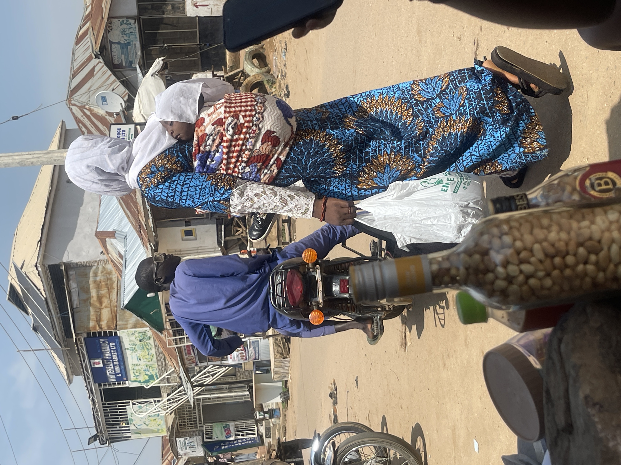 The image captures a street scene in Abuja, Nigeria, likely during the daytime given the bright sunlight and clear sky. The primary subjects are two people on a motorcycle. A man in a blue tunic is driving the motorcycle, and a woman in a vibrant blue and gold patterned dress and a white headscarf is seated behind him. She is carrying a white plastic bag and appears to be resting her head, perhaps due to fatigue or the movement of the motorcycle. The woman's dress is adorned with a striking floral or tree-like pattern.
The setting is a bustling urban environment with buildings that appear to be shops or market stalls. The architecture suggests a local, perhaps somewhat informal, commercial area with corrugated metal roofs and signs advertising businesses. One visible sign reads "WHITEHEART PHARMACY & MINI-MARKET LTD," providing a specific location cue. Other signs are partially visible or obscured.
In the foreground, out of focus, are two glass bottles. One is filled with what appear to be nuts or seeds, and the other has a dark liquid and a label with the letter "B" prominently displayed. These objects suggest a commercial stall or a vendor's display. The overall atmosphere is one of everyday life in a busy Nigerian city, with a focus on the movement and interaction of people within their environment. The image is oriented vertically, suggesting it was taken with a phone held in portrait mode.