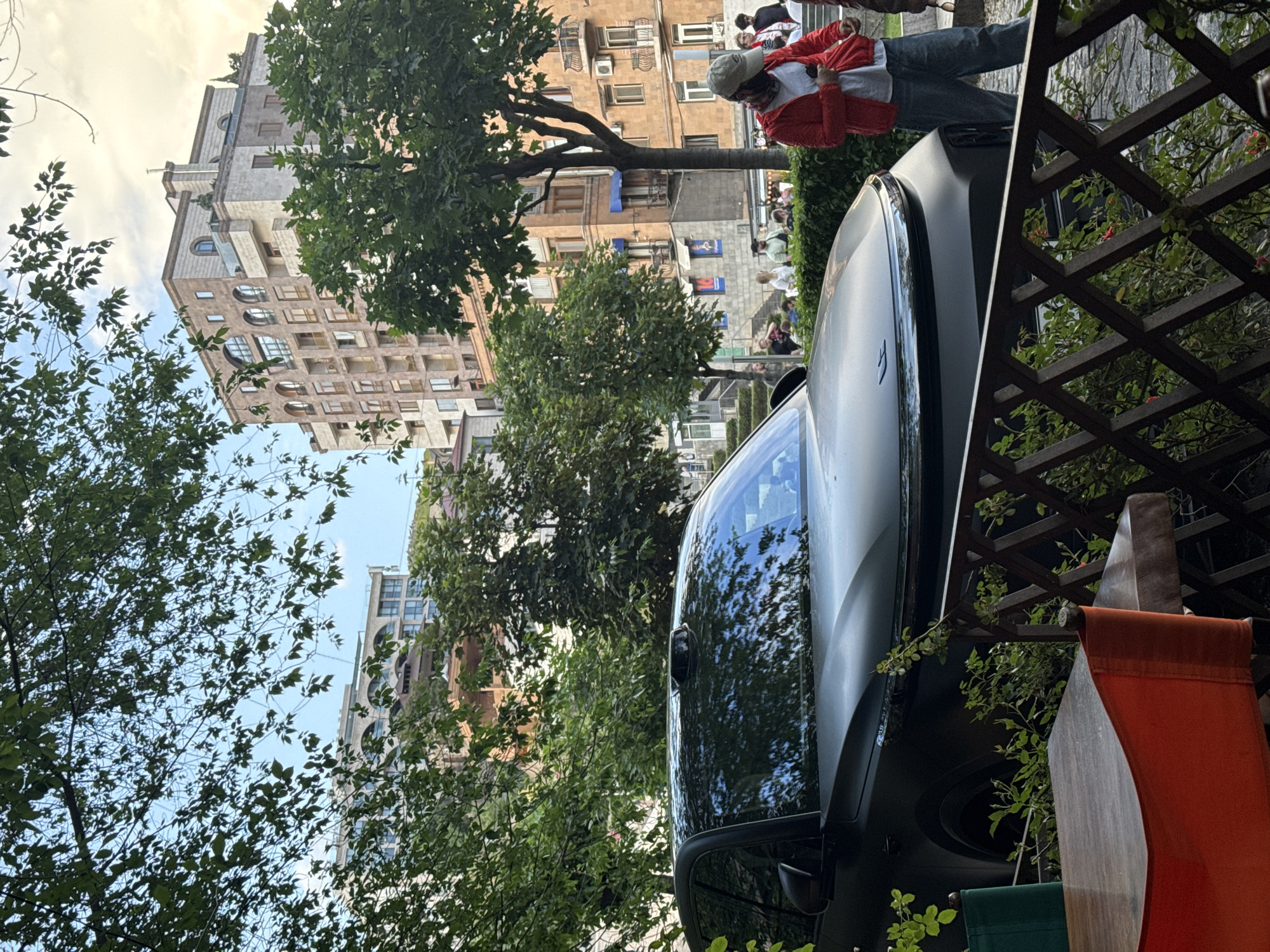 The image shows an urban scene in Yerevan, Armenia. A modern silver car dominates the right side of the frame, parked beside a wooden trellis with climbing plants and a red fabric hanging. The buildings on the left showcase a mix of architectural styles, with one prominent beige building featuring rounded windows. A crowd of people is visible in the background on the right, where a man in a red jacket stands near a tree. The sky is cloudy, and trees with green foliage are scattered throughout the scene.