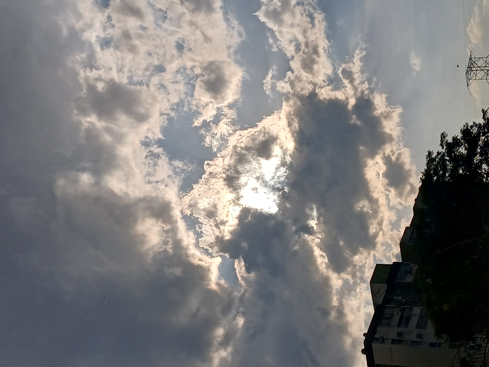 The image displays a dramatic sky dominated by a dynamic interplay of bright white and dark grey cumulus clouds. The sun intensely breaks through a central clearing in the clouds, creating powerful highlights and sun rays that illuminate the edges of the cloud formations. The lighting suggests a daytime scene, possibly late afternoon or early morning, characterized by strong contrasts between sunlit areas and shadowed cloud masses.
In the lower right section of the frame, the dark silhouette of a multi-story building is visible, featuring several air conditioning units on its side. Adjacent to the building, dense, dark green tree foliage extends into the scene. Towards the upper right, a portion of a metal power line tower structure can be seen against a lighter, clearer patch of sky. Small, dark specks, likely birds, are intermittently visible against the brighter sky and clouds.
The photograph is taken from a low-angle perspective, emphasizing the vastness and intricate details of the cloudy sky. The overall impression is one of natural beauty and atmospheric grandeur, with a hint of an urban or semi-urban setting in the foreground elements. No visible text is present. The context provided indicates the image was taken in Ciudad Guayana, Venezuela.