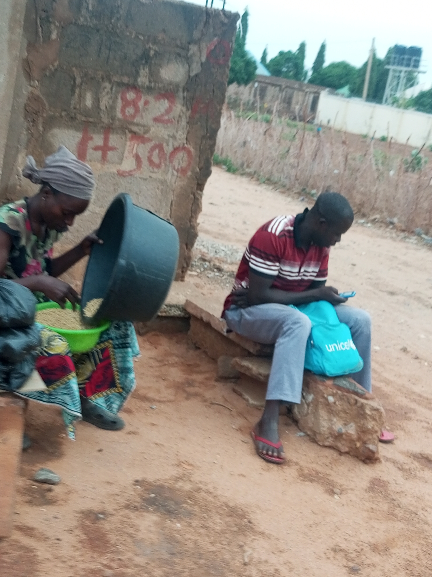 The image shows two individuals outdoors in what appears to be the city of Tempure, Nigeria.
To the left, a woman is seated on a low ledge or step near a weathered wall with faded red markings, possibly numbers. She is pouring grains from a large black bucket into a green bowl. She wears a headwrap and patterned clothing.
To the right, a man is seated on a rough stone or brick structure, looking at a mobile phone. A bright blue UNICEF bag rests between him and the seat. He wears a red and white striped shirt, gray pants, and flip-flops.
The ground is sandy and appears to be outdoors. Background shows a fence, greenery, and buildings in the distance.