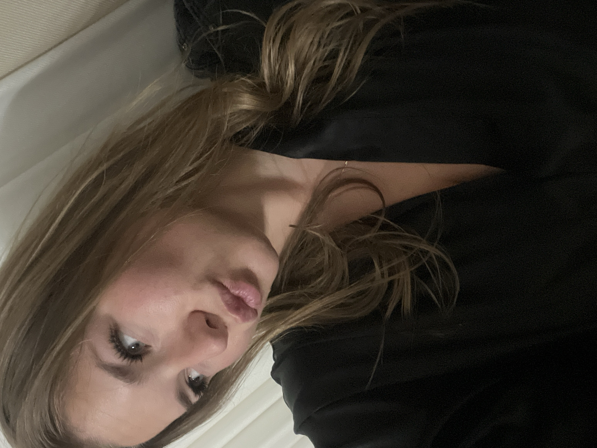 A woman with light brown hair is seen in a close-up shot. She has fair skin, light eyes, and full lips. She is wearing a black top. The background appears to be a white surface, possibly a bed or a couch. The photograph is slightly rotated. The setting appears to be indoors with limited light. There is no information about the location in the city of Budva, Montenegro.
