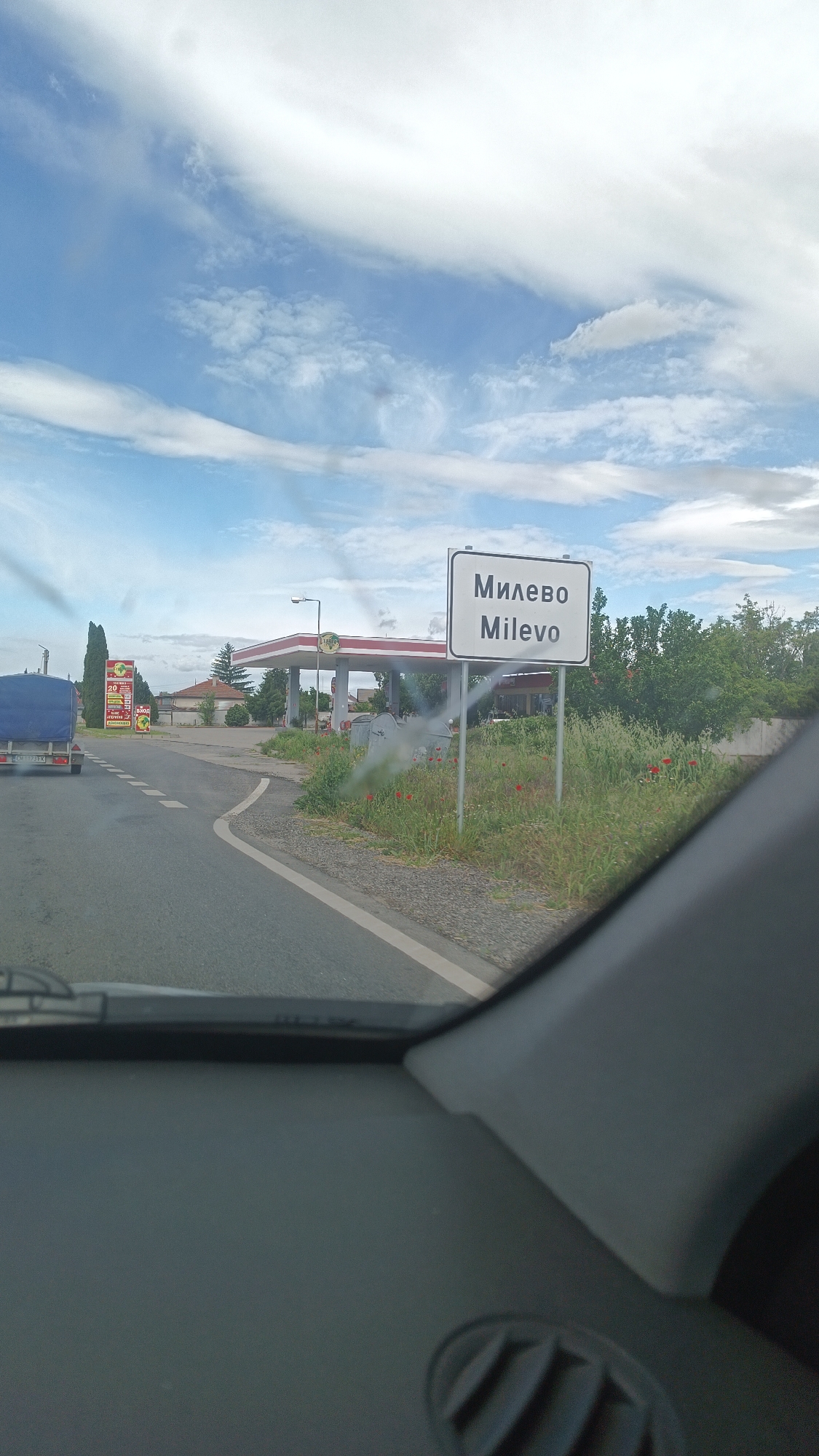 The image, taken from inside a vehicle, shows the road sign for the village of Milevo, Bulgaria. The sign is white with black Cyrillic and Latin text. In the background, there's a gas station with a red and white canopy. To the left of the road is a trailer hitched to a vehicle and a sign advertising prices in Bulgarian. The sky is mostly cloudy. The dashboard of the vehicle is visible in the foreground. The road is asphalt with white lane markings. Grass and wildflowers grow along the roadside near the sign.