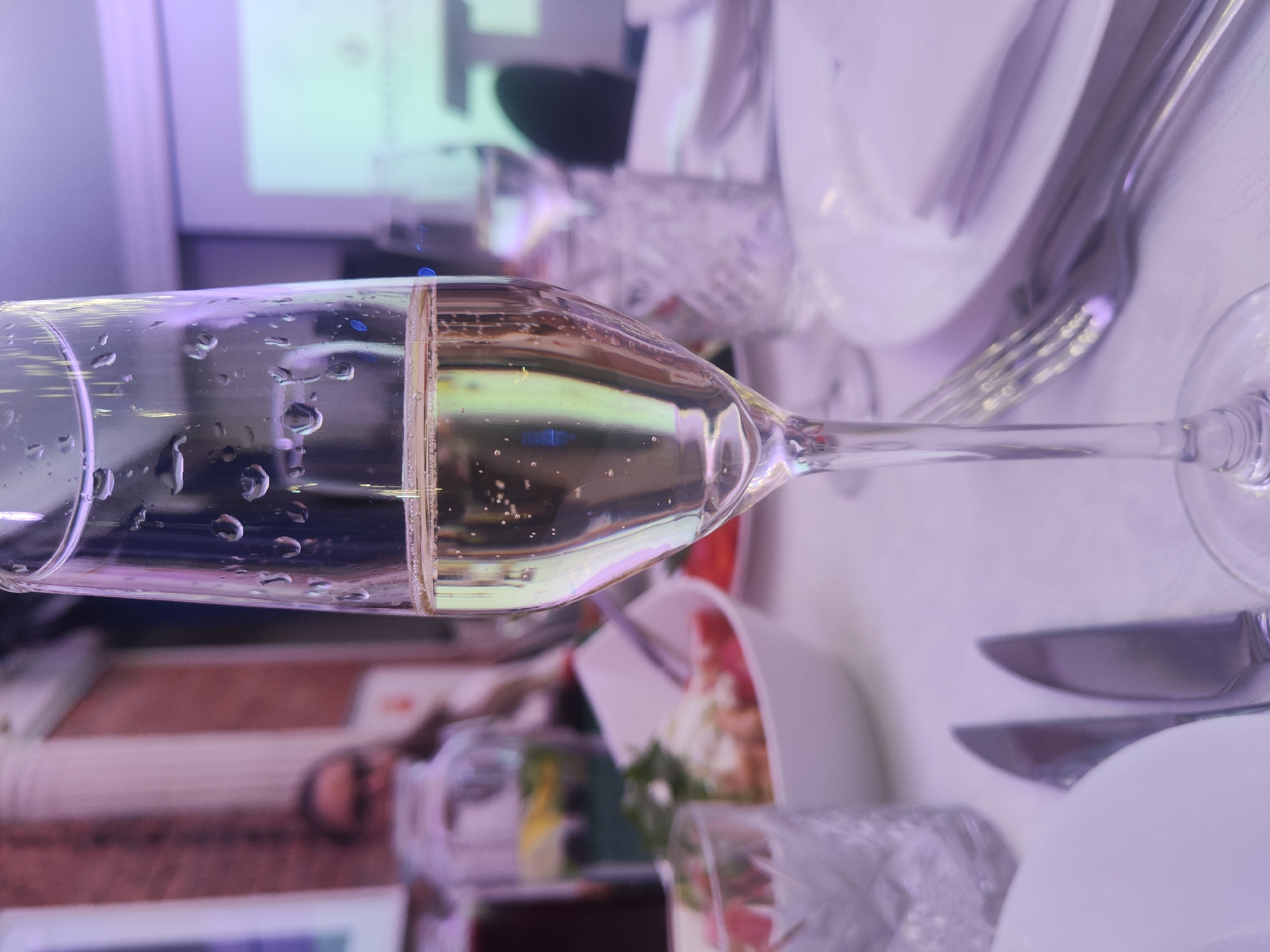 A close-up shot focuses on a champagne flute filled with a light-colored beverage, its surface adorned with condensation droplets. The flute is placed on a white tablecloth, with cutlery and plates visible in the foreground and background, suggesting a formal dining setting. In the blurred background, a projector screen displays an image, indicating a gathering or event. To the left, a woman is partially visible, wearing glasses and looking towards the front. The overall ambiance is dimly lit, with purple and white hues dominating the scene. This image was taken at a restaurant in Saint Petersburg, Russia.