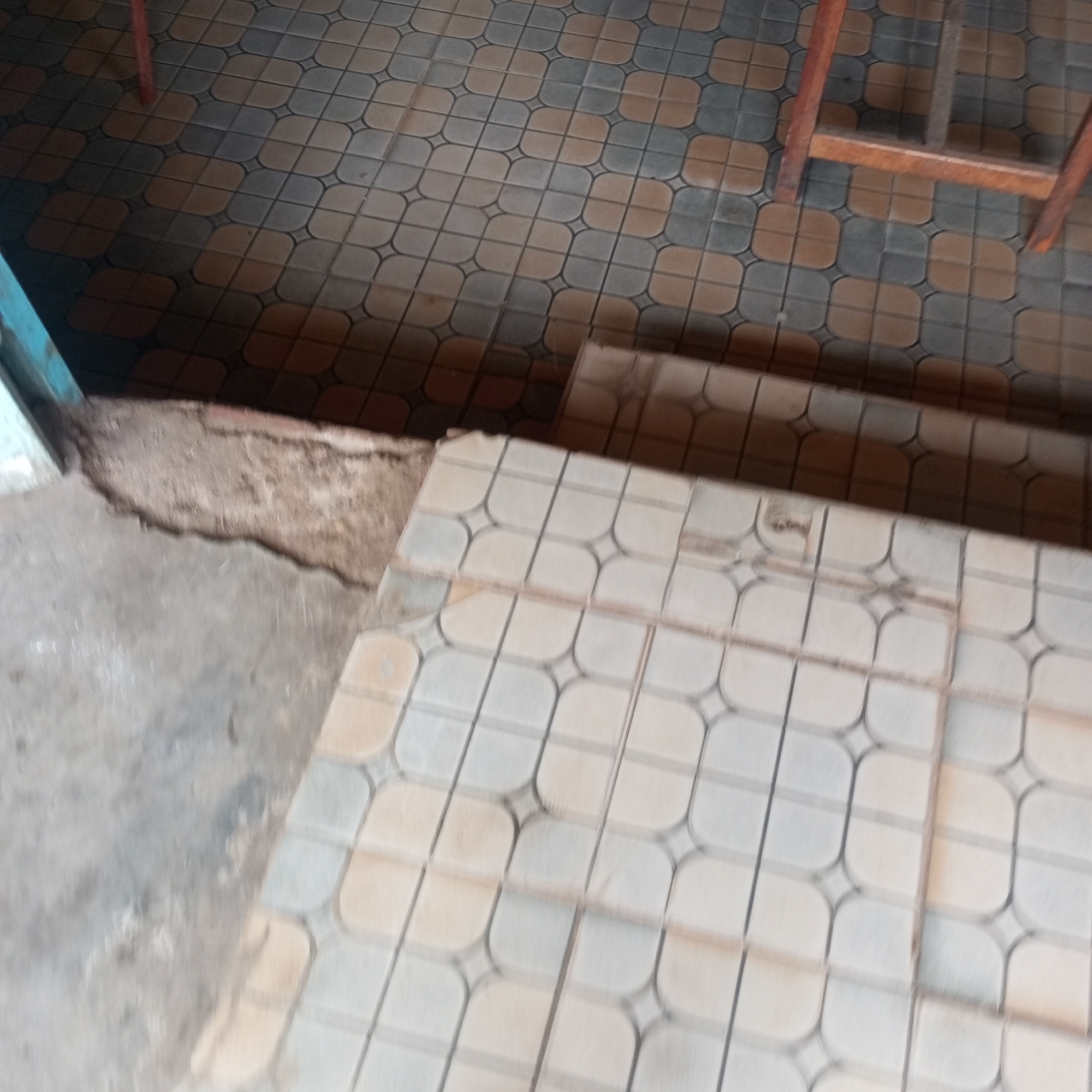 The image shows a portion of a tiled floor with two different patterns. One pattern features alternating brown and gray tiles, while the other has light gray tiles with a darker gray grid. A wooden structure is visible in the upper right corner. A small step with the light gray tiles leads down to a rough, concrete area near what appears to be a doorframe on the left. The location is Sheywe ward, Kenya.