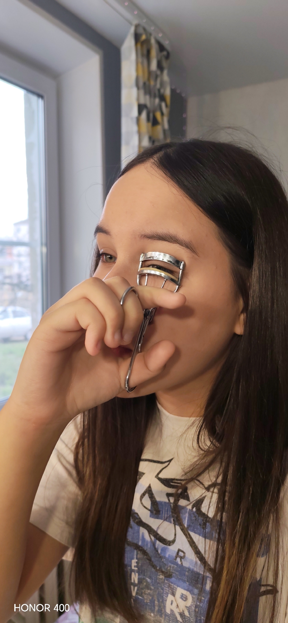 A young woman with long brown hair is shown from the chest up in an indoor setting. She is holding a silver eyelash curler up to her left eye, her fingers visible around the curler's handles. She is wearing a white t-shirt with a blue and black design on the front.
A window is visible to the left, showing an overcast day outside with buildings and a car in the distance. A patterned curtain hangs on the right. The phone used to take the photo is identified as an "HONOR 400" at the bottom left. The location can be assumed to be somewhere in Russia but it is not identified.