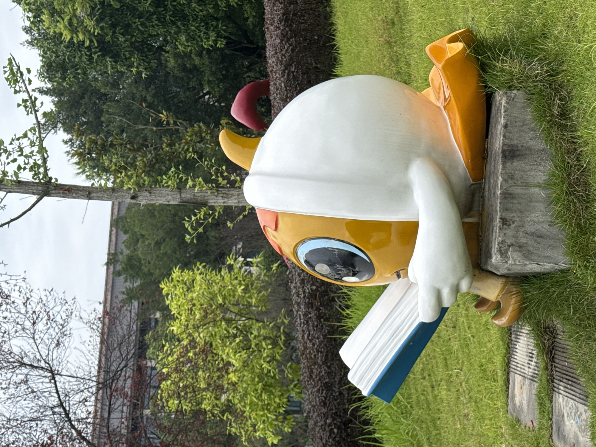 This is a medium shot, eye-level, outdoor photograph of a cartoonish statue of a yellow, round character reading a book. The statue is situated on a grassy knoll in front of a manicured hedge and a building with a tiled roof. The statue has a large, blue-rimmed eye with a black pupil, and a white, cowl-like covering over its head. It is wearing orange shoes and has a red tuft on its head. The book it is reading is open and appears to have blue covers. The background consists of green trees and a cloudy sky. The location of this statue is in the city of Jiangjin, China, specifically at a local library.