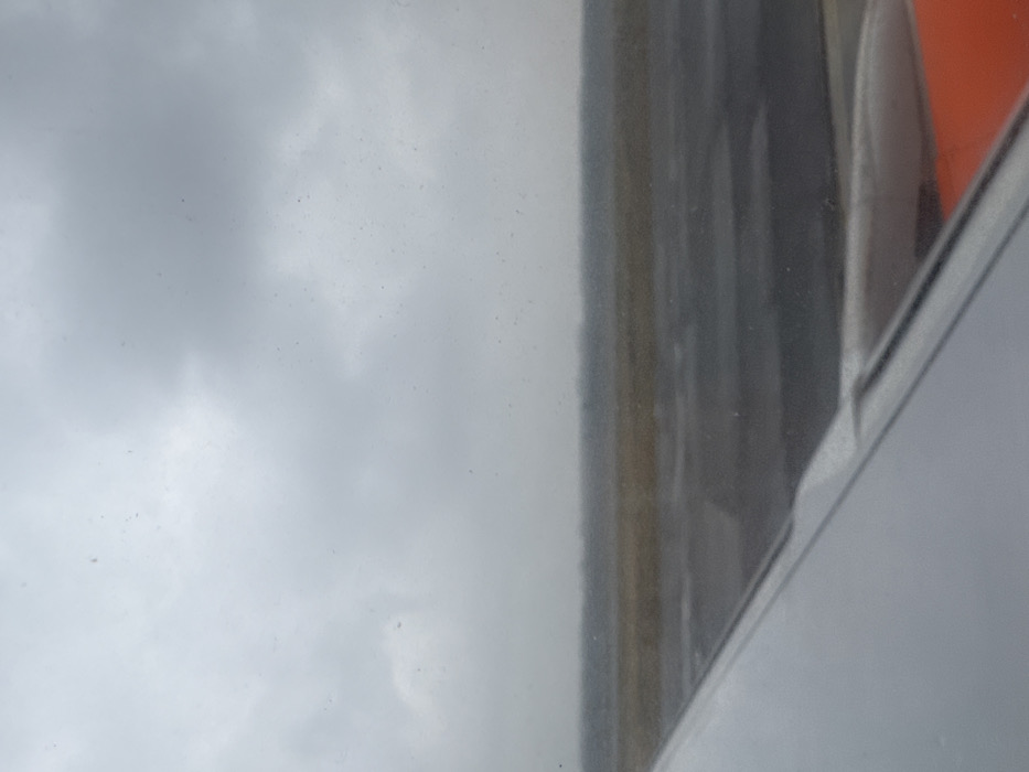 The image presents a forensic view from inside an aircraft at Milan Malpensa Airport (MXP) in Somma Lombardo, Italy. The lower left quadrant displays a portion of a grey and white aircraft wing. Below the wing's trailing edge, in the bottom right, an orange component, likely part of an engine cowling, is visible. The midground consists of a dark grey asphalt or concrete runway surface, appearing wet and reflective, suggesting recent precipitation. Adjacent to the runway is a strip of muted brown, possibly dry ground or vegetation. The horizon line is indistinct, with a faint, hazy line of dark, blurred forms in the far background, indicative of distant landscape or structures. The sky is entirely overcast, dominated by uniform grey clouds, indicating low visibility conditions. Small water droplets or specks are scattered across the aircraft windowpane. No people are visible. The scene captures the static presence of an aircraft on the tarmac under a cloudy, possibly rainy, sky.