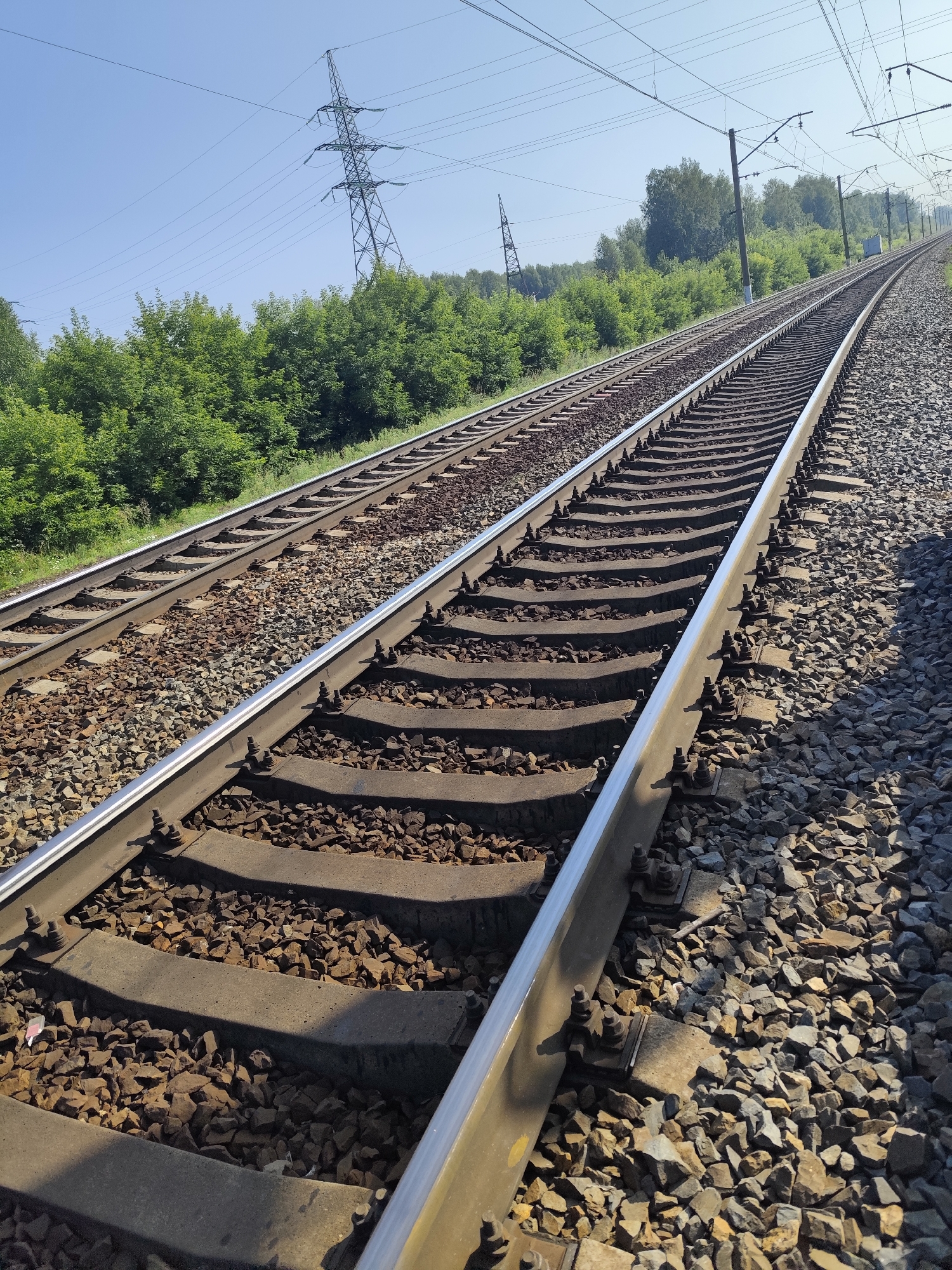 The image shows a section of railroad tracks in the city of Barlak, Russia. The tracks are made of steel rails laid on concrete sleepers, all resting on a bed of crushed stone ballast. The tracks stretch into the distance, curving slightly to the right. On the left side of the image, another set of parallel tracks is visible. The area is bordered by a dense green forest on the left and slopes upwards towards more trees in the background. Power lines and tall, lattice-like electricity pylons are present in the sky above, indicating the presence of an overhead power transmission network. The sky is clear and blue, suggesting a sunny day. No people or vehicles are visible in the frame. The perspective is from ground level, looking along the tracks.
