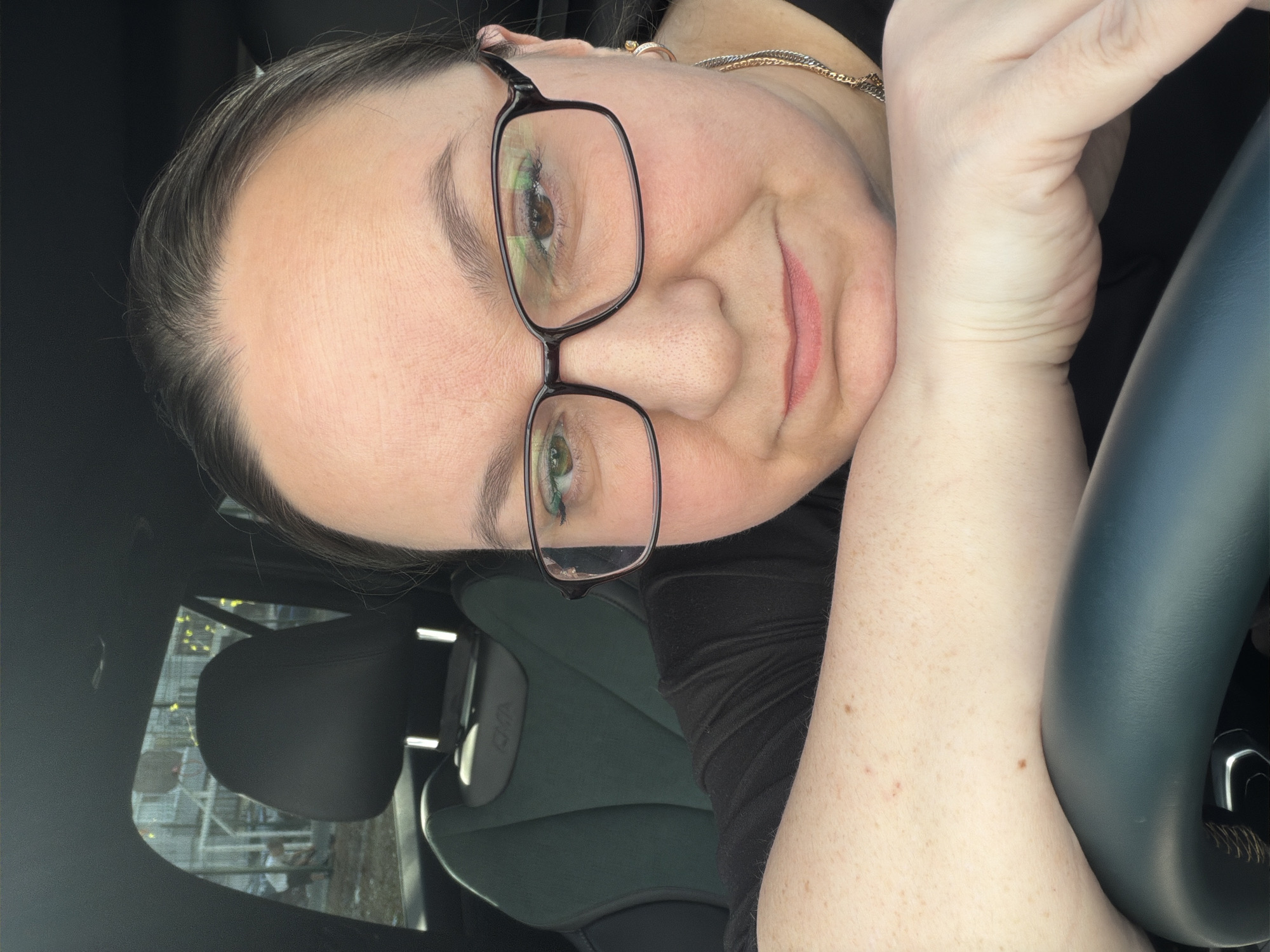 A woman with fair skin and dark hair pulled back is pictured in a close-up selfie, likely taken inside a car. She is wearing black-framed glasses, a dark shirt, and a gold necklace. Her left arm is resting on the steering wheel. A car seat and headrest are visible in the background. The scene outside the car window shows an indistinct structure with string lights or decorations. Based on the request, this is presumed to be in Chaikovsky, Russia.