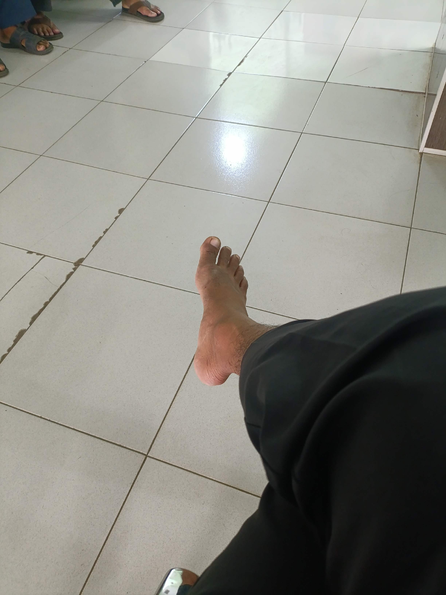 This is a first-person perspective image of a bare foot resting on a tiled floor. The foot is dark-skinned with visible toes and some hair on the ankle and lower leg. The person is wearing dark pants. The floor is made of light-colored square tiles with visible grout lines. There are some faint marks and smudges on the tiles. In the background, on the left side of the image, the feet of other individuals are visible, wearing sandals and flip-flops, suggesting a public waiting area or gathering. The overall environment appears to be indoors with a smooth, reflective floor. This scene could be located in Banyumas, Indonesia.