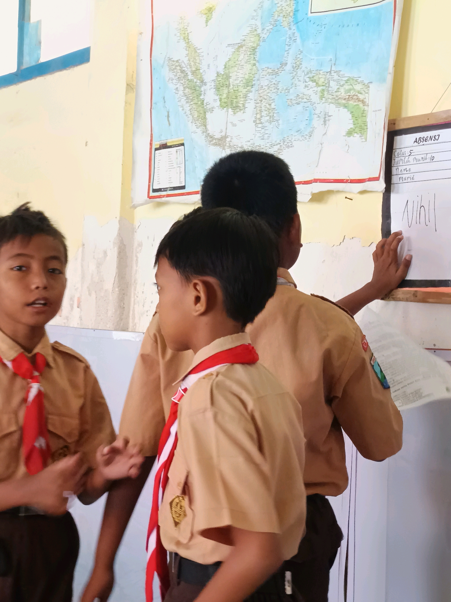 The image captures a scene in a classroom in Labuhan, Indonesia, likely a school. Three young boys, dressed in brown school uniforms with red and white scout scarves, are the central subjects. A world map is displayed on the wall behind them, and a whiteboard is also visible, with some handwritten text that appears to be an attendance list, including "ABSENSI," "Kelas 5," "Jumlah Murid: 10," "Nama," "Murid," and the word "Nihil."
The boy on the left is looking directly at the camera with his mouth slightly open, engaged in what appears to be a conversation or explanation. The boy in the center, facing away from the camera, is also engaged in the activity, possibly pointing at or interacting with the whiteboard. The third boy, on the right, is mostly obscured by the other two but his arm is extended towards the whiteboard, and he is holding a piece of paper. The classroom environment is suggested by the map and whiteboard, and the general appearance of the wall indicates a well-used educational space. The lighting suggests it is daytime.