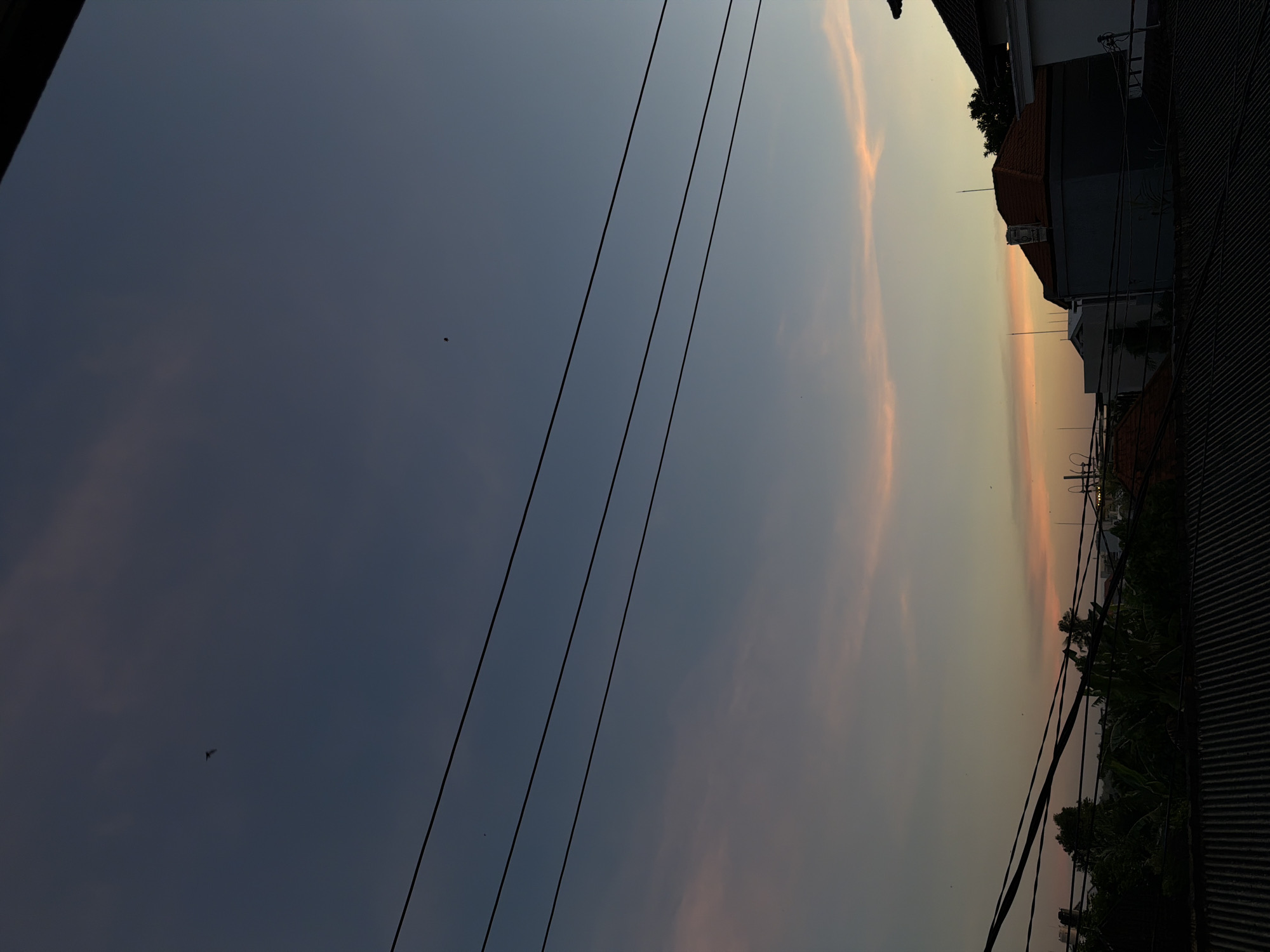 The image shows a late afternoon or early evening sky in North Kuta, Indonesia. The sky transitions from a dark blue-grey on the left to a light orange-yellow hue near the horizon on the right. Several electrical or telephone wires stretch across the frame from top left to lower right, partially obscuring the view. The silhouettes of buildings and rooftops, characteristic of an urban or suburban area, are visible on the right, along with some green foliage which could be trees or shrubs. There is a small, dark shape of a bird flying high up in the sky.