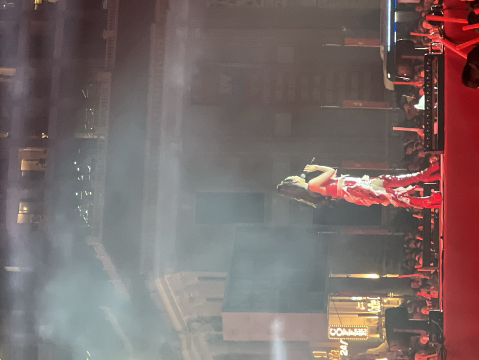 The image displays a nighttime outdoor concert scene in Nha Trang, Vietnam. A female performer, characterized by long dark hair styled in a high ponytail, occupies the center-right of the frame on an elevated red stage. She wears a red two-piece outfit, consisting of a crop top and a ruffled skirt with white accents, complemented by tall, red, platform boots. She holds a microphone to her mouth with her right hand, suggesting vocal performance.
The stage is brightly illuminated by spotlights, generating visible beams of light through atmospheric haze or smoke. Below the stage, a dense crowd is visible, with numerous individuals holding glowing red light sticks. Stage lighting equipment is positioned in front of the audience area.
In the background, a large, multi-story building with ornate architecture and a distinctive roofline is visible. Illuminated windows indicate activity within the structure. On the lower left facade of this building, a brightly lit commercial sign displays "24/7" and "Coffee" in English, suggesting a retail or hospitality establishment. Other less distinct signs are visible further right on the building. The overall scene indicates a public event in an urban environment.