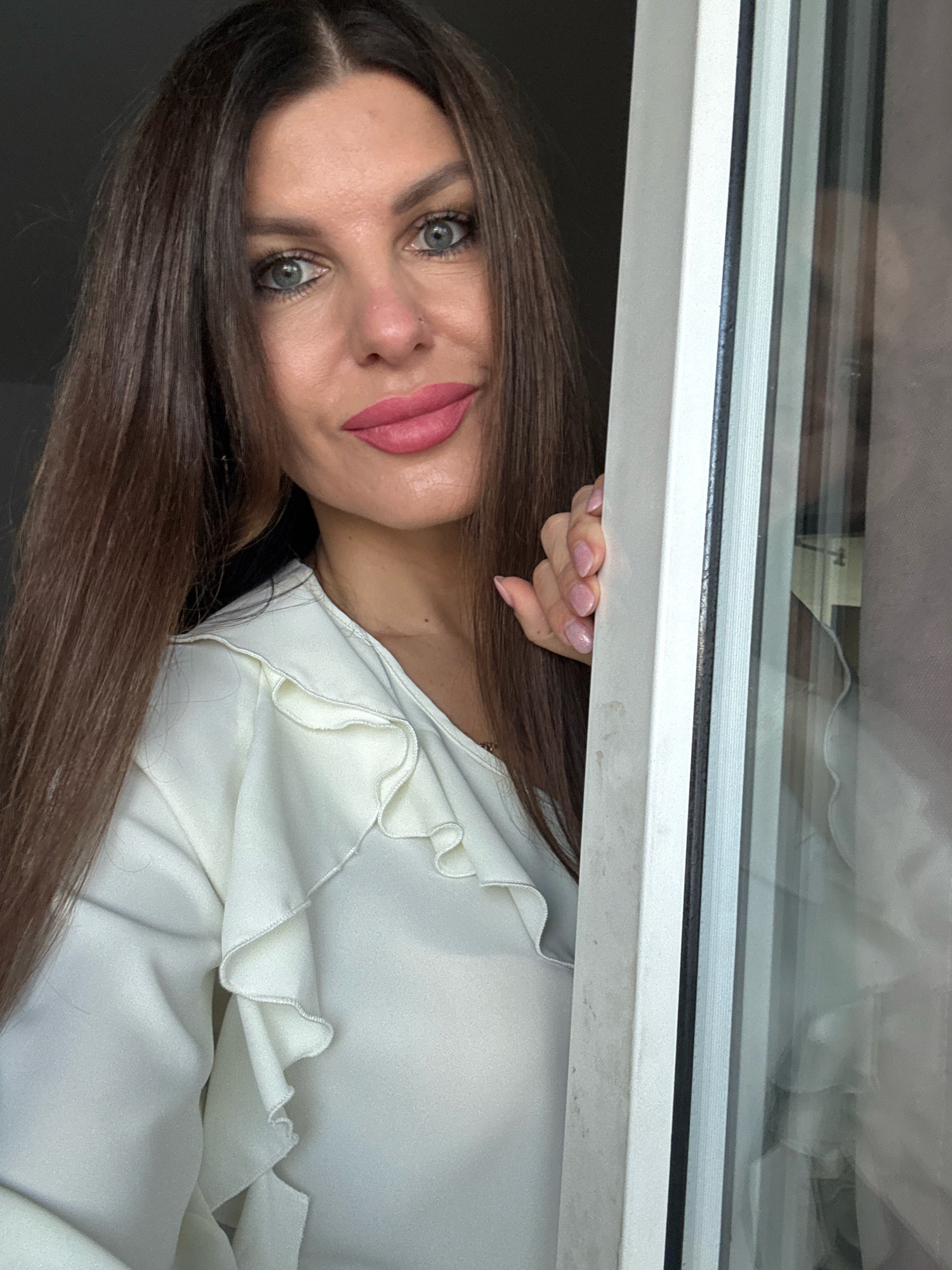 A woman with long, dark brown hair and blue eyes is looking out from behind a window frame. She is wearing a white blouse with ruffles. Her right hand is resting on the window frame. The background is dark, suggesting an indoor setting. There is no indication of the specific location within the city of Chaikovsky, Russia.