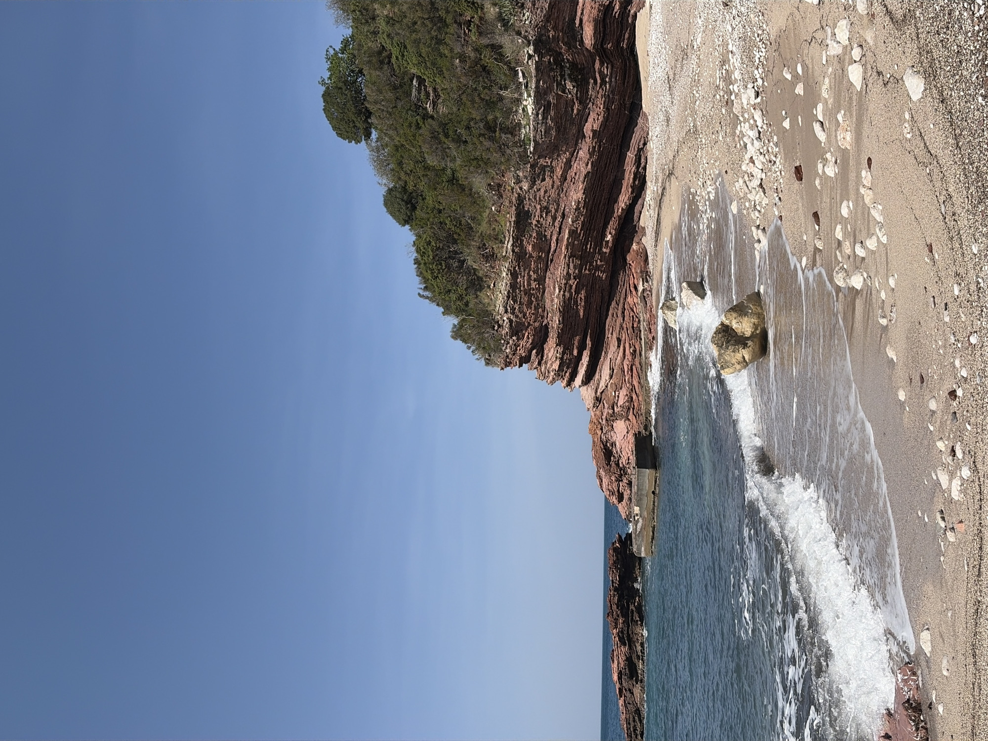 The image displays a serene coastal landscape, rotated 90 degrees clockwise from its natural orientation, showing a steep, rugged cliff, a beach, and the sea under a clear blue sky.
On the left side of the frame (when correctly viewed in landscape), a prominent, reddish-brown, layered rock cliff rises dramatically, covered with lush green vegetation, including trees and bushes, particularly dense at the top. At the base of this cliff, a narrow strip of sandy and pebbly beach extends towards the right and bottom of the frame. The beach is composed of light-colored sand mixed with numerous white and darker pebbles and rocks of varying sizes.
The right side and bottom of the image are dominated by the clear, blue-green sea. Gentle waves are seen breaking on the shore, creating white foam and ripples that wash over the beach. Several larger rocks are visible both partially submerged in the water and resting on the sandy shore. A distinct, flat rock formation or possibly a man-made structure juts out into the water from the base of the cliff on the lower left.
The upper portion of the scene is filled with a bright, cloudless blue sky, indicating clear weather. The lighting suggests it is daytime, likely late morning or early afternoon, with the sun illuminating the scene clearly. There are no people, animals, or visible text in the image. The overall impression is one of natural beauty and tranquility.