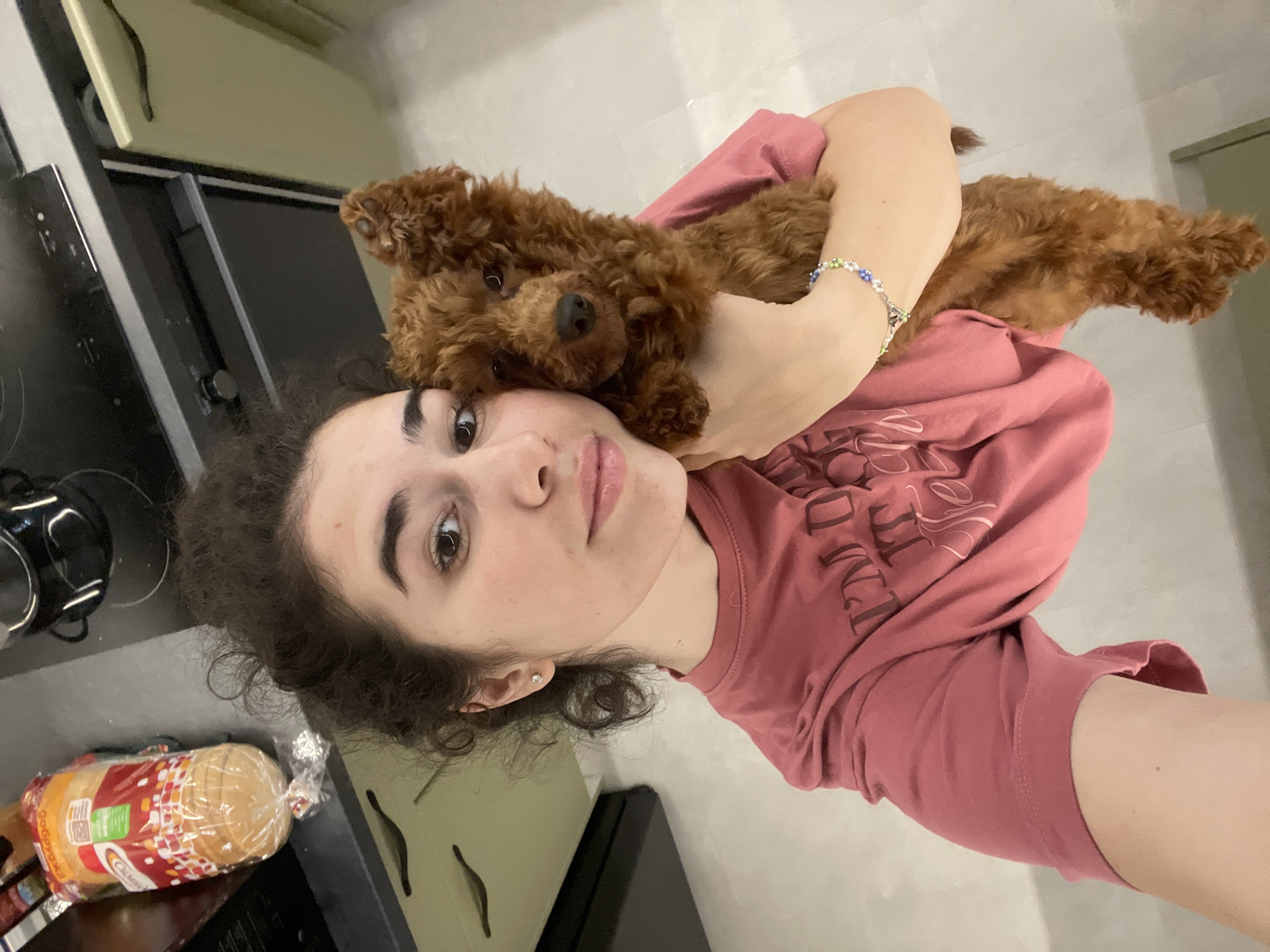 A young woman is holding a small, brown poodle. She is wearing a pink t-shirt and a beaded bracelet. The woman has curly, dark hair and is wearing a small earring. She is looking at the camera with a neutral expression. The poodle is nestled against her chest. The background includes a kitchen counter with a stove, a loaf of bread in a plastic bag, and various kitchen cabinets. The specific location in Sofia, Bulgaria, cannot be determined from the image.
