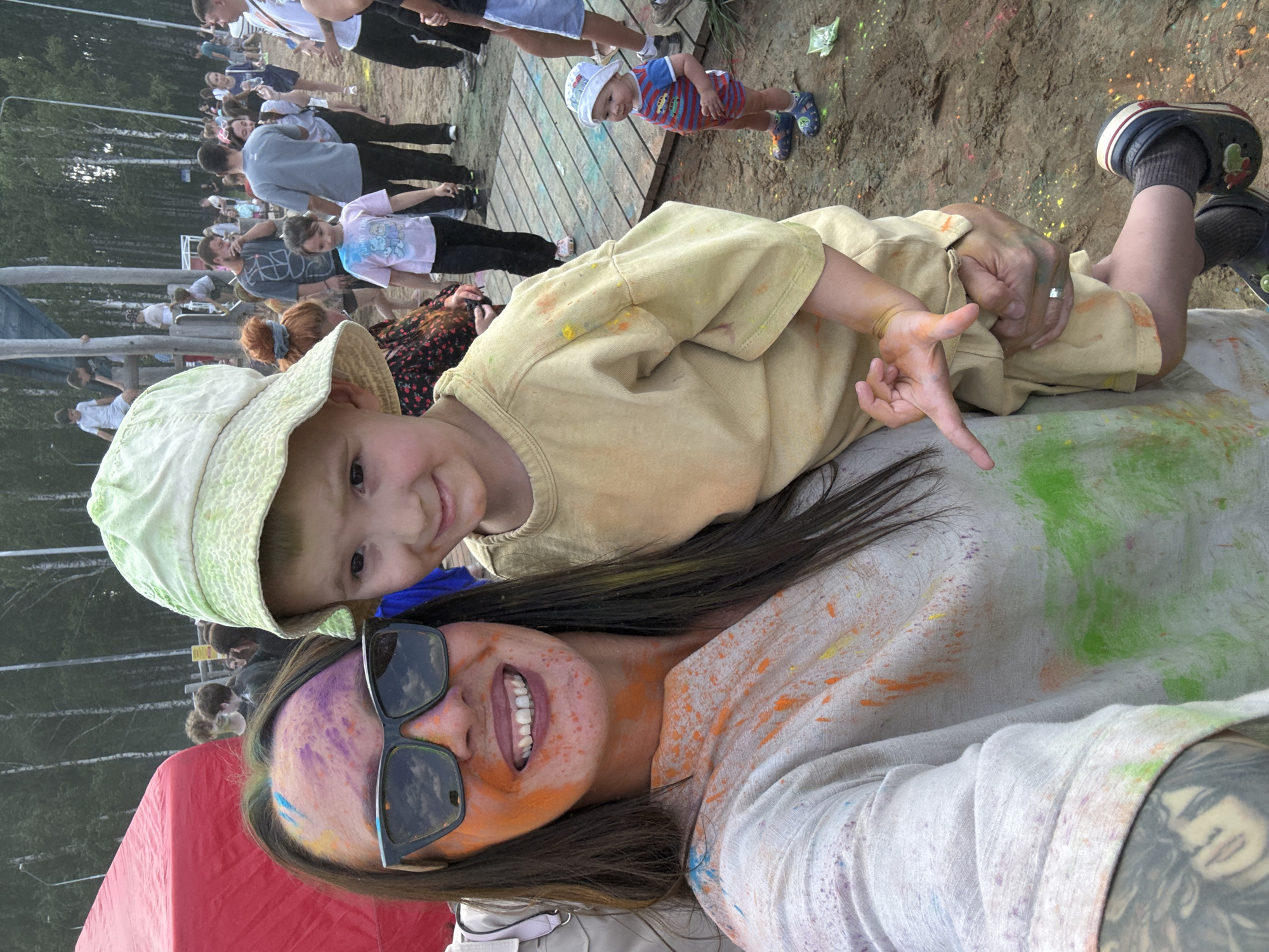 A group of people, including adults and children, are gathered at an outdoor event, possibly a festival, in Chaikovsky, Russia. The ground is sandy and scattered with colored powder, suggesting a color-themed activity like Holi. A woman in the foreground is holding a child, and both have colored powder on their faces and clothing, indicating participation in the event. The woman is wearing sunglasses and a light-colored garment, while the child is wearing a yellow t-shirt and a light-colored bucket hat. In the background, other people are seen socializing and participating in activities. The environment appears to be a park or open space with trees lining the background.