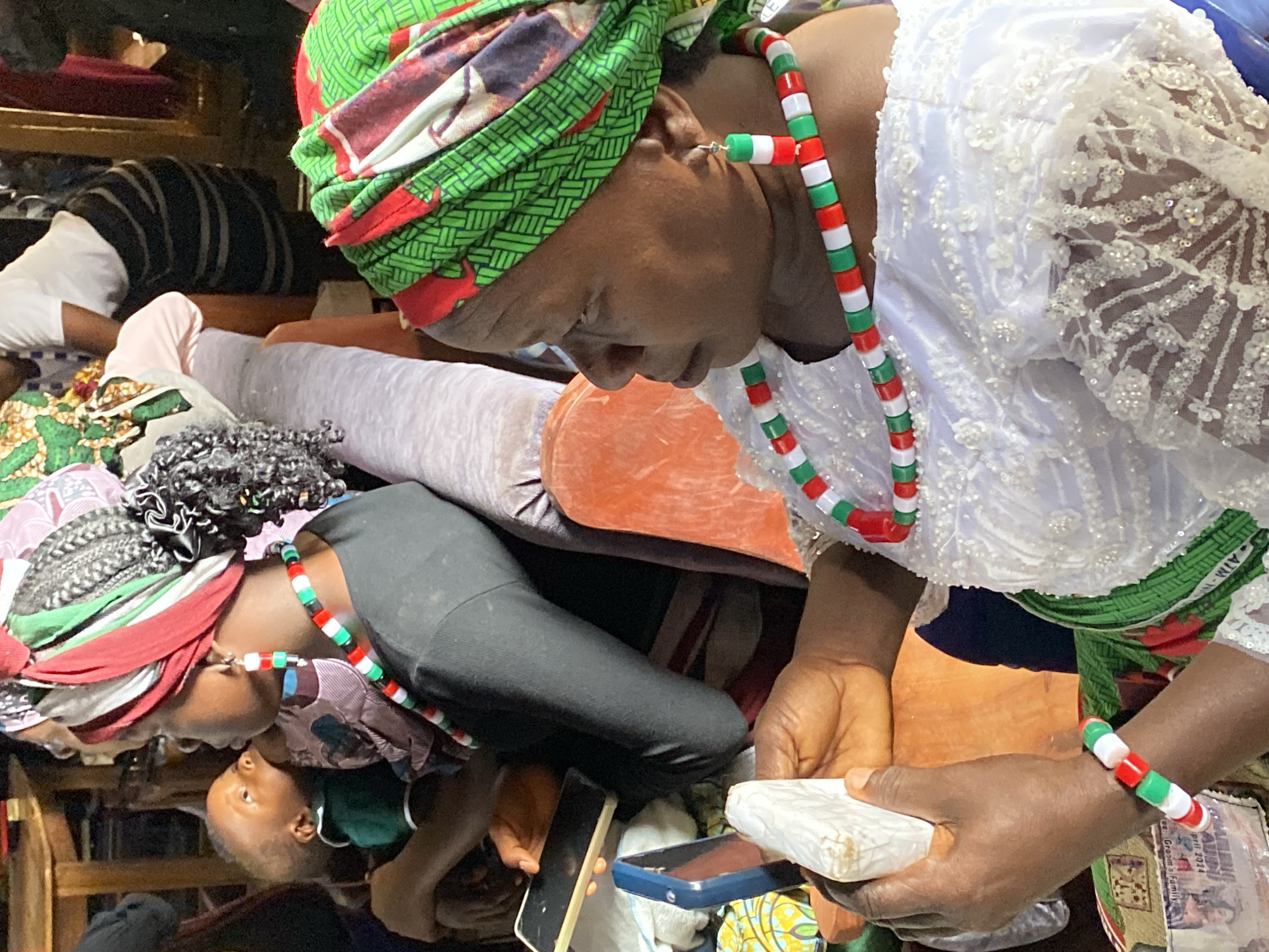 The image shows an indoor scene with multiple individuals. An adult woman wearing a green and red headwrap and a white lace top is holding a white rectangular object in her hand. She is also wearing a necklace and a bracelet with red, white, and green beads. A child is facing upwards towards another adult wearing similar attire. Several mobile phones and other personal items are scattered around. The setting appears to be a room with various objects and fabrics. It is impossible to confirm the location as Durimi, Nigeria from this image alone.