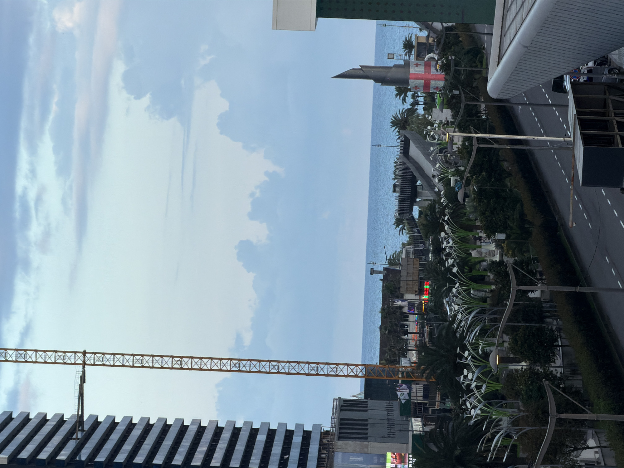 The image captures a view of Batumi, Georgia, showcasing a coastal city with a mix of modern architecture and public spaces. In the foreground, a multi-lane road runs alongside a pedestrian area featuring unique, stylized lamp posts resembling plants or trees, with some illuminated. Beyond this, there is a waterfront promenade with palm trees and various structures, including a large, abstract sculpture adorned with the Georgian flag. The sea is visible in the background, meeting a sky filled with scattered clouds. To the left, a tall, unfinished skyscraper is under construction, with a yellow construction crane attached to its side. Across the street, other buildings and lights suggest an urban environment. The overall impression is a developing cityscape with recreational areas adjacent to the sea.