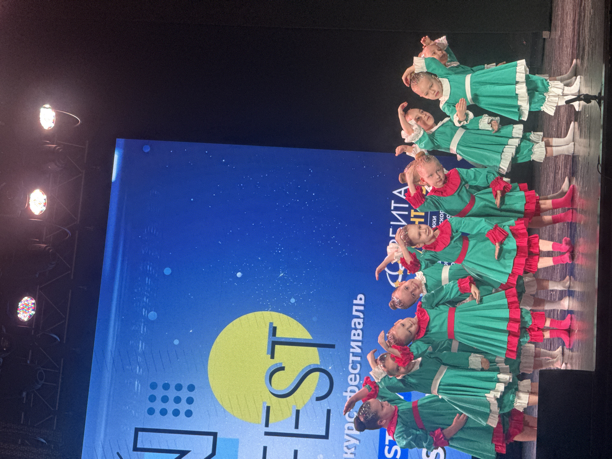 The image depicts a group of young girls performing a dance routine on a stage. They are dressed in matching green costumes with red or white ruffles and wearing matching socks. They appear to be in the middle of a coordinated movement. Behind them is a large screen with the word "FEST" in large letters and the Russian words for "course festival." The image was taken at an event located in Yekaterinburg, Russia. Stage lighting fixtures are visible above the stage and to the sides of the image.