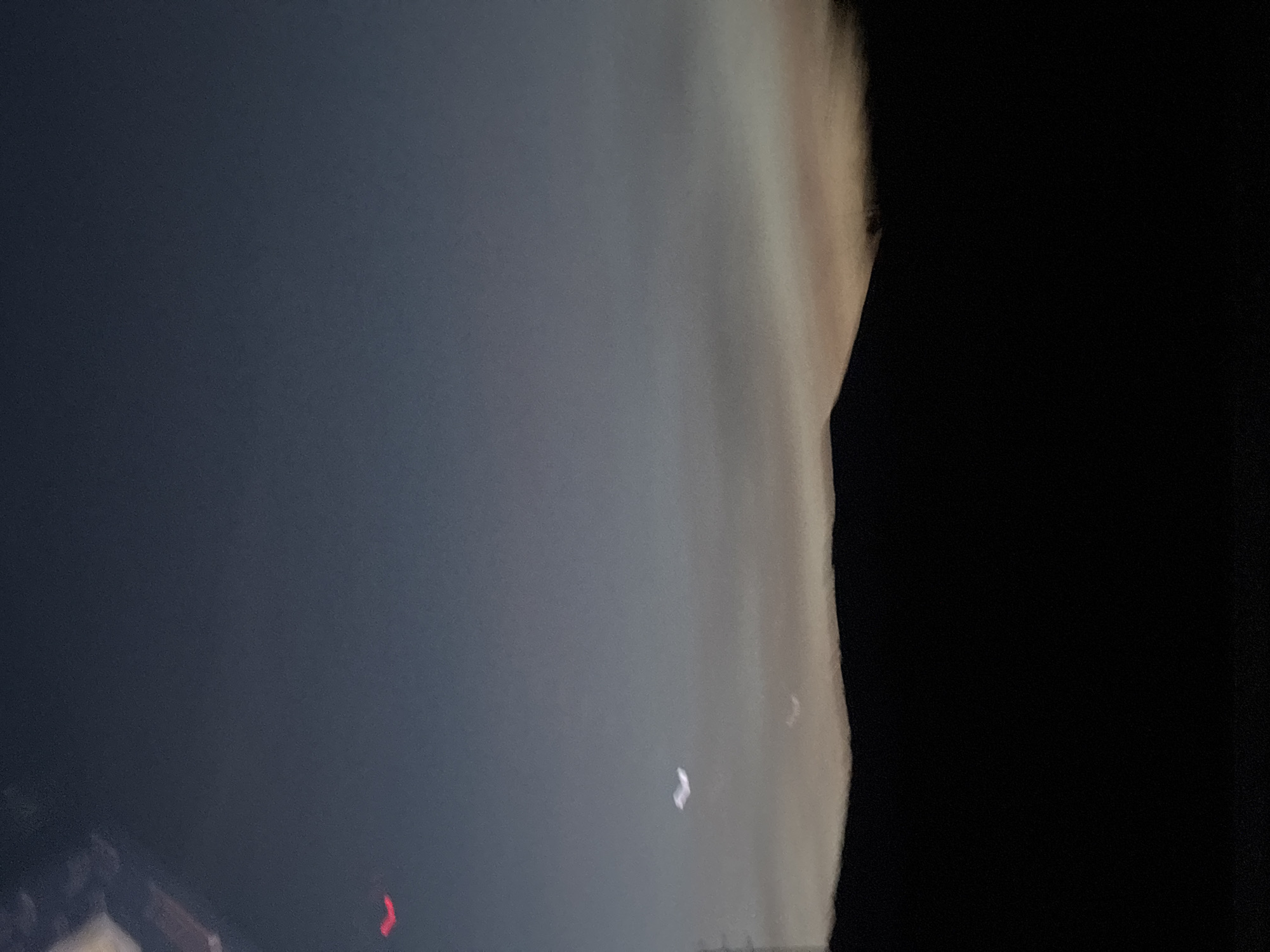 The image shows a dimly lit, overcast night sky over what appears to be an urban area. There is a dark, undefined structure or mass, possibly a building or foliage, silhouetted on the right side of the image. A faint, red light source is visible at the lower left, along with what might be parts of a building or infrastructure. There are also some streaks or distortions of light, possibly clouds or atmospheric conditions in the sky, with a pale, yellow hue near the right. There are also some indistinct bright spots in the sky, possibly stars or streetlights. The location is described as Mirandela, Portugal. The image appears to be a low-quality photo taken under poor lighting conditions.