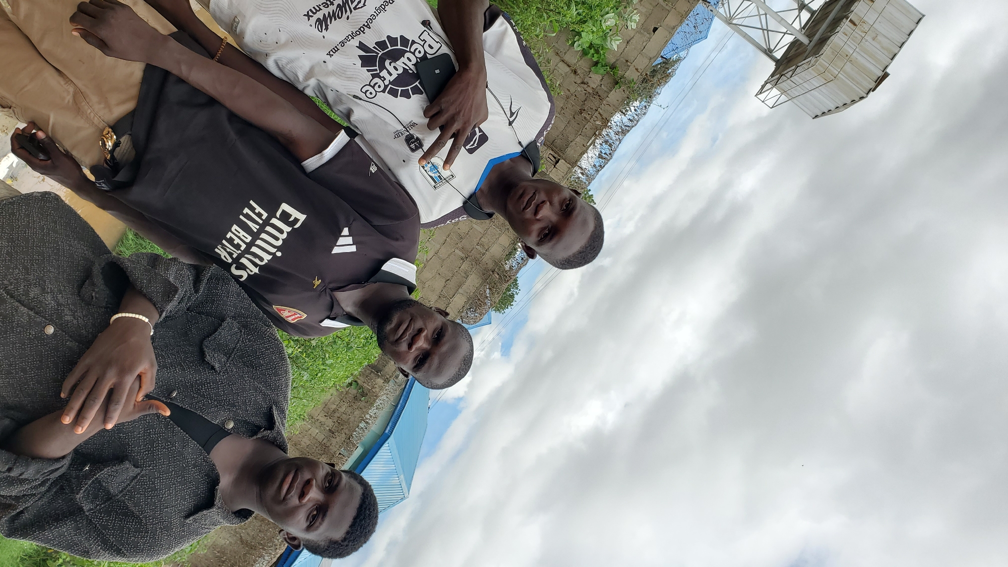 Three young men are standing in an outdoor urban environment, likely in Jalingo, Nigeria, under a cloudy sky. The man on the left, wearing a white and black shirt with blue trim and the word "Pedigree" printed on it, is making a peace sign with his right hand and holds a black smartphone in his left. The man in the middle is wearing a black polo shirt with white accents and the text "Emirates Fly Better." He is looking directly at the camera. The man on the right is wearing a dark, textured jacket over a black shirt and is also looking towards the camera. They are standing in front of a brick wall with barbed wire on top, and lush green vegetation is visible. In the background, a large metal water tower is visible on the left, and a blue-roofed building is present on the right. The ground appears to be muddy or wet in the foreground.